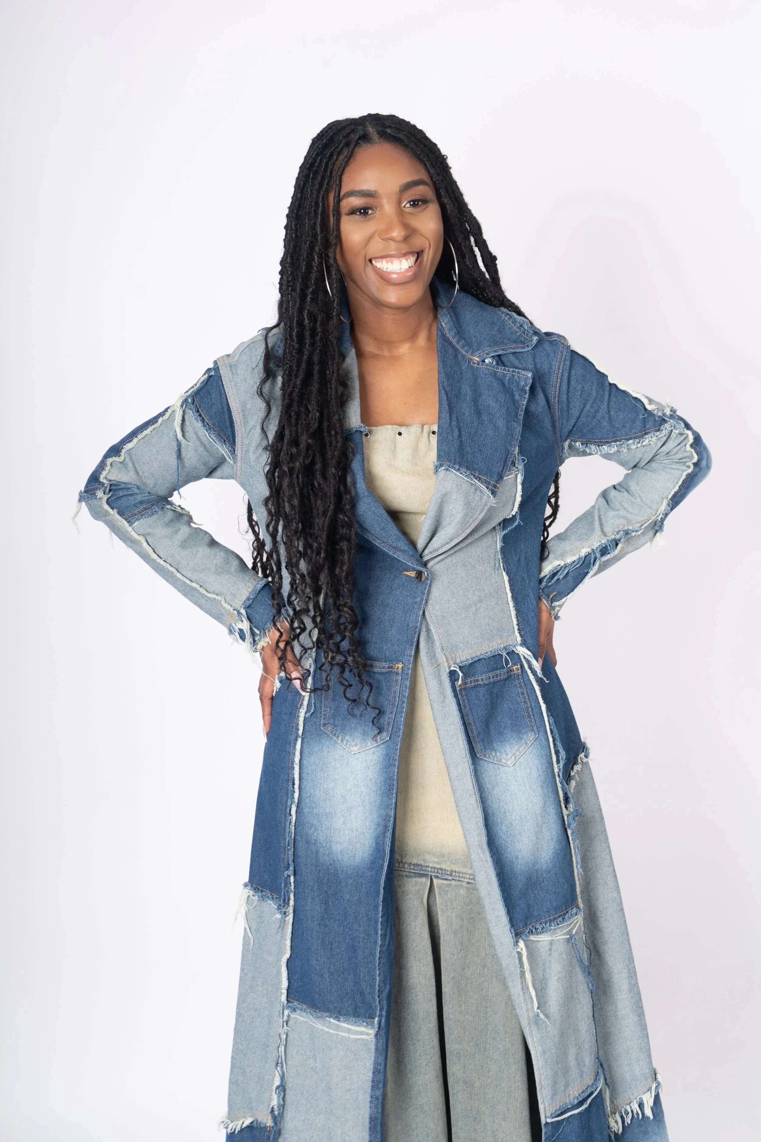 A woman with long curly hair smiling and standing with hands on hips, wearing a patchwork denim long coat over a beige dress, against a plain white background.