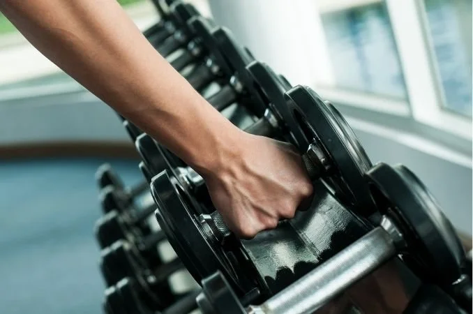 Close-up of a person's hand gripping a black dumbbell in a gym near large windows