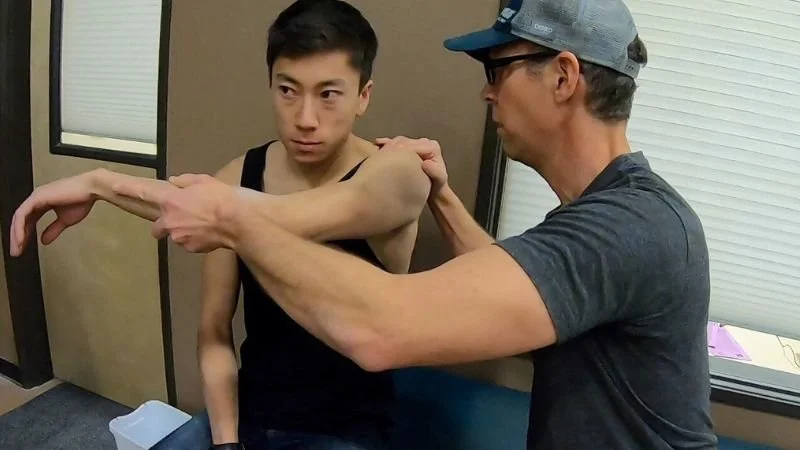 A sports therapist and coach is helping a young male climber stretch his arm while seated in a climbing gym room with beige walls and a window.