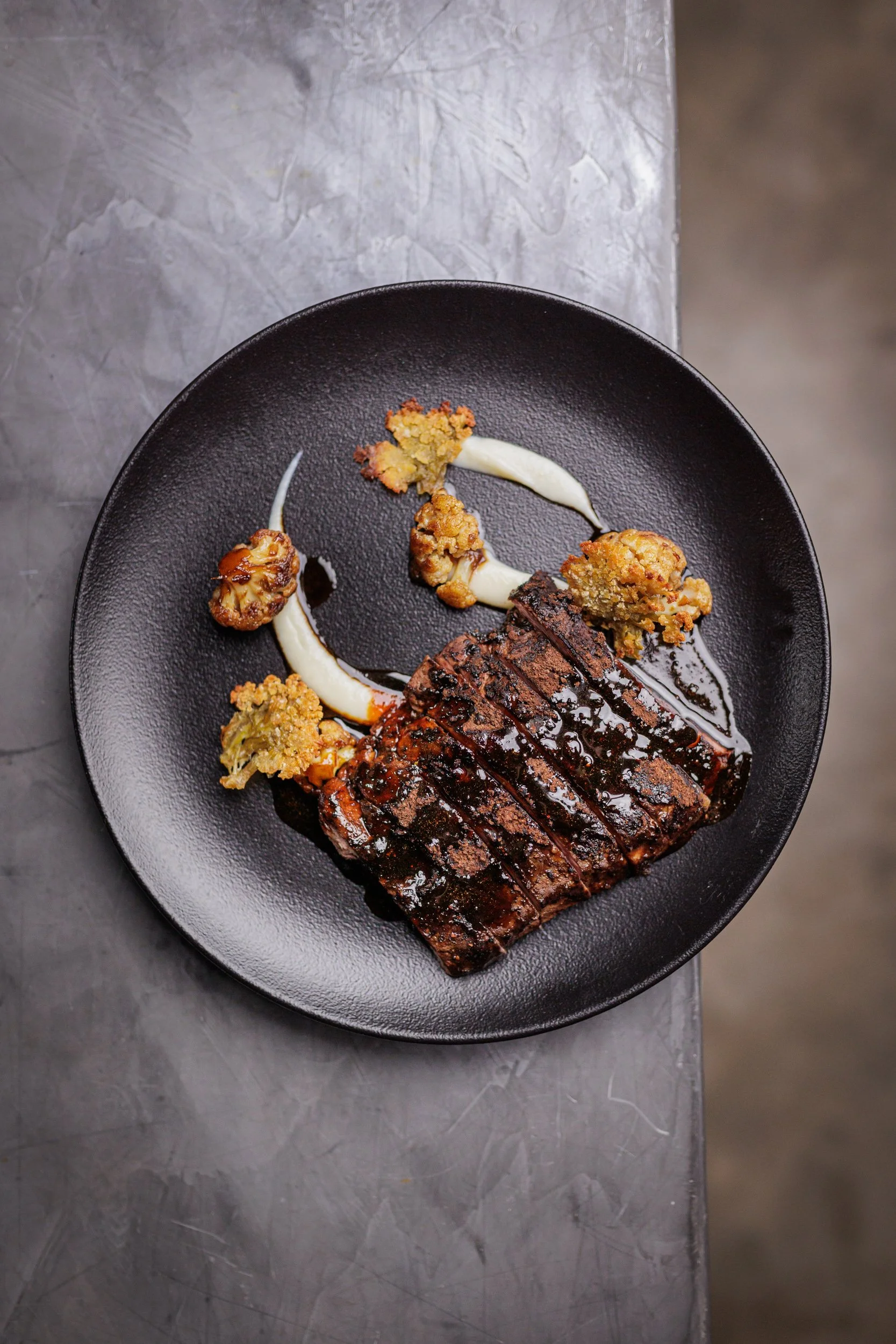 Pechuga de pollo a la parrilla con salsa, acompañada de vegetales fritos y una crema blanca, en un plato negro.
