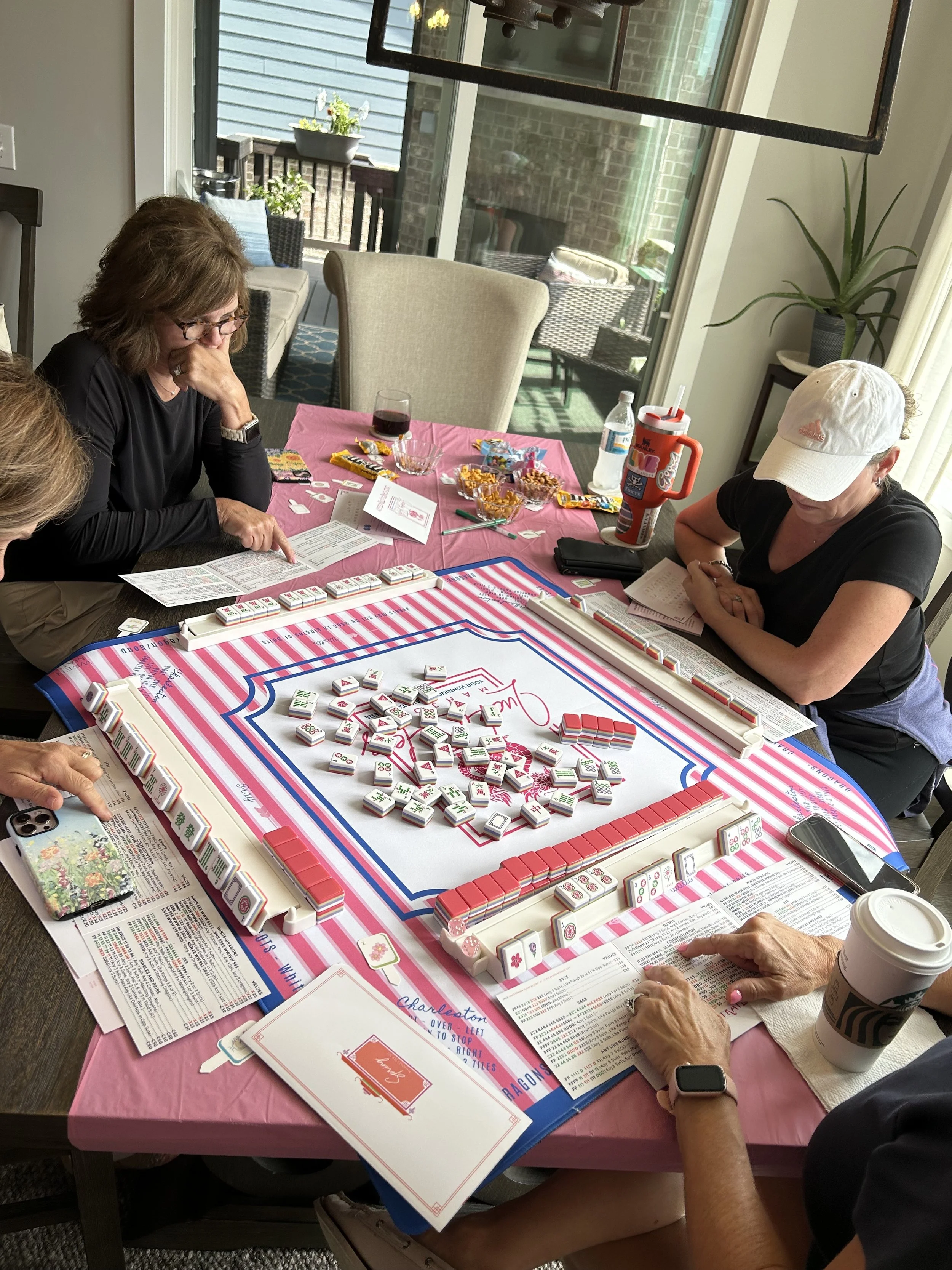 People playing Mahjong around a table with mahjong tiles, snacks, and drinks in a bright room with large windows.