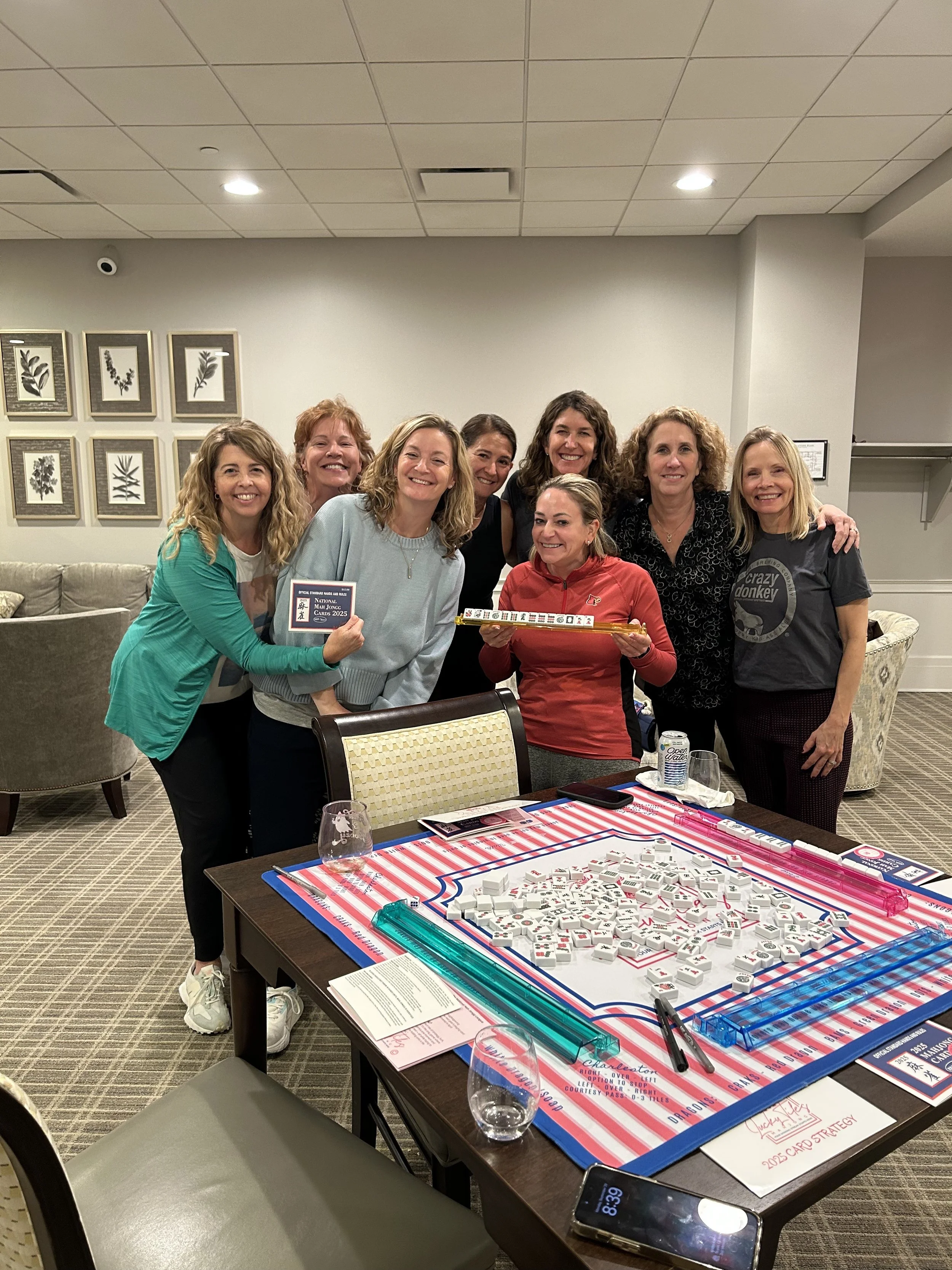 Group of women smiling and celebrating around a table with Mahjong tiles, cards, and glasses, in a room with framed artwork on the wall.