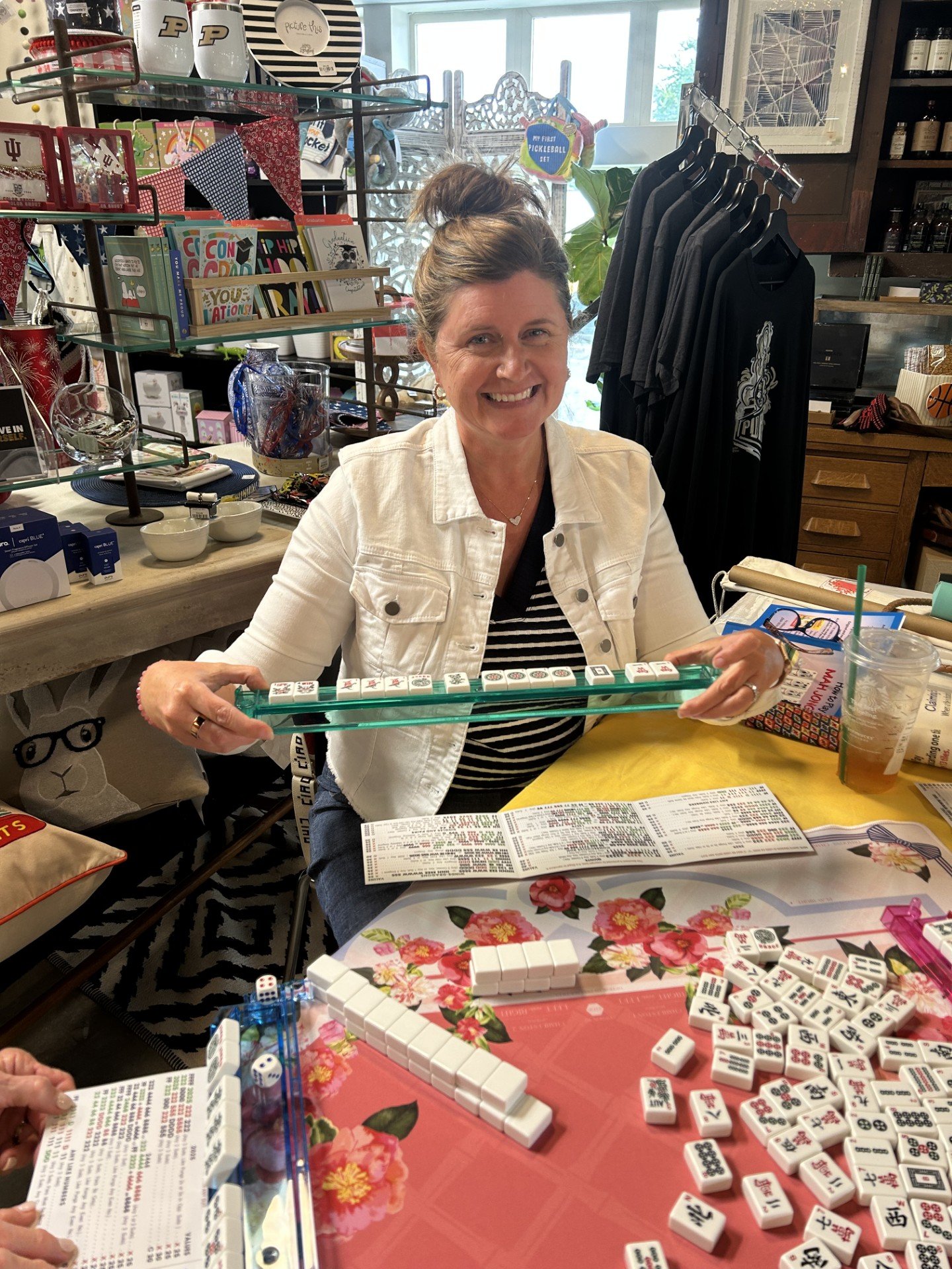 A woman smiling and sitting at a table playing mahjong, holding a mahjong tile rack. The table has mahjong tiles, dice, and score sheets on it. The background shows a store with shelves of books, mugs, and clothing.