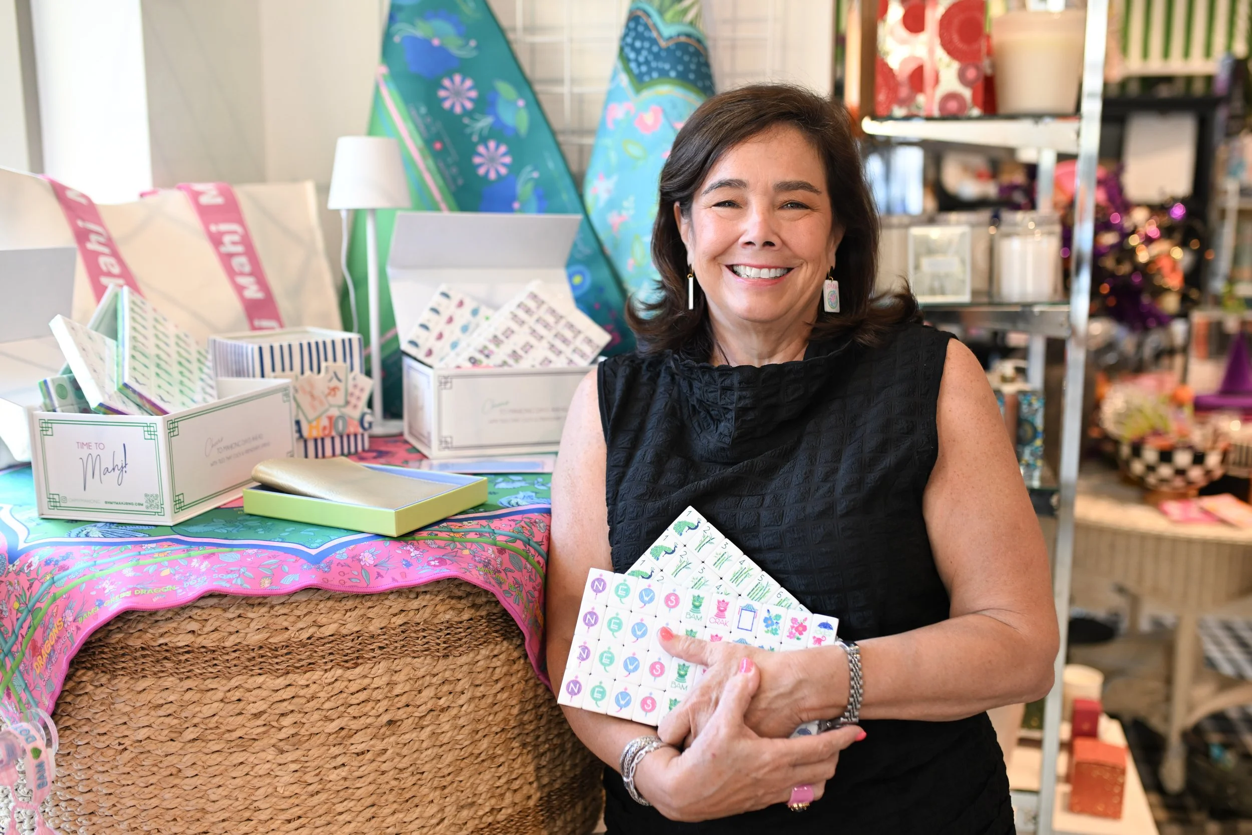 A smiling woman in a black sleeveless top holding colorful stickers in a craft store with various craft supplies, boxes, and decorations on display.