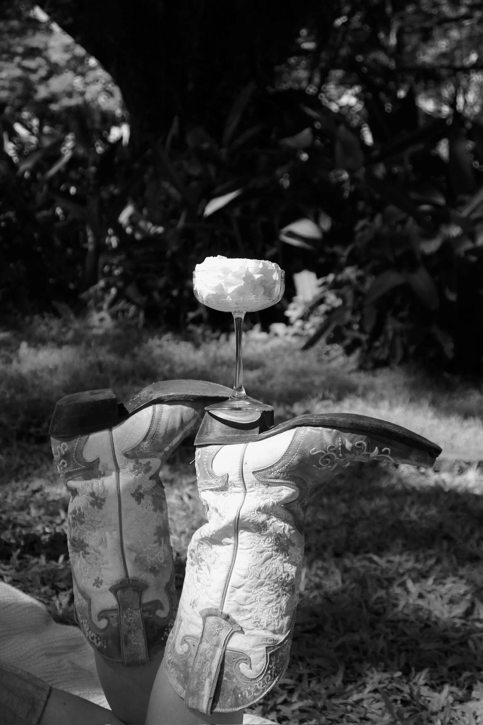 A black and white photo showing a pair of cowboy boots with a stemmed glass balanced on top. The glass contains a frothy beverage. Background features trees and grass.