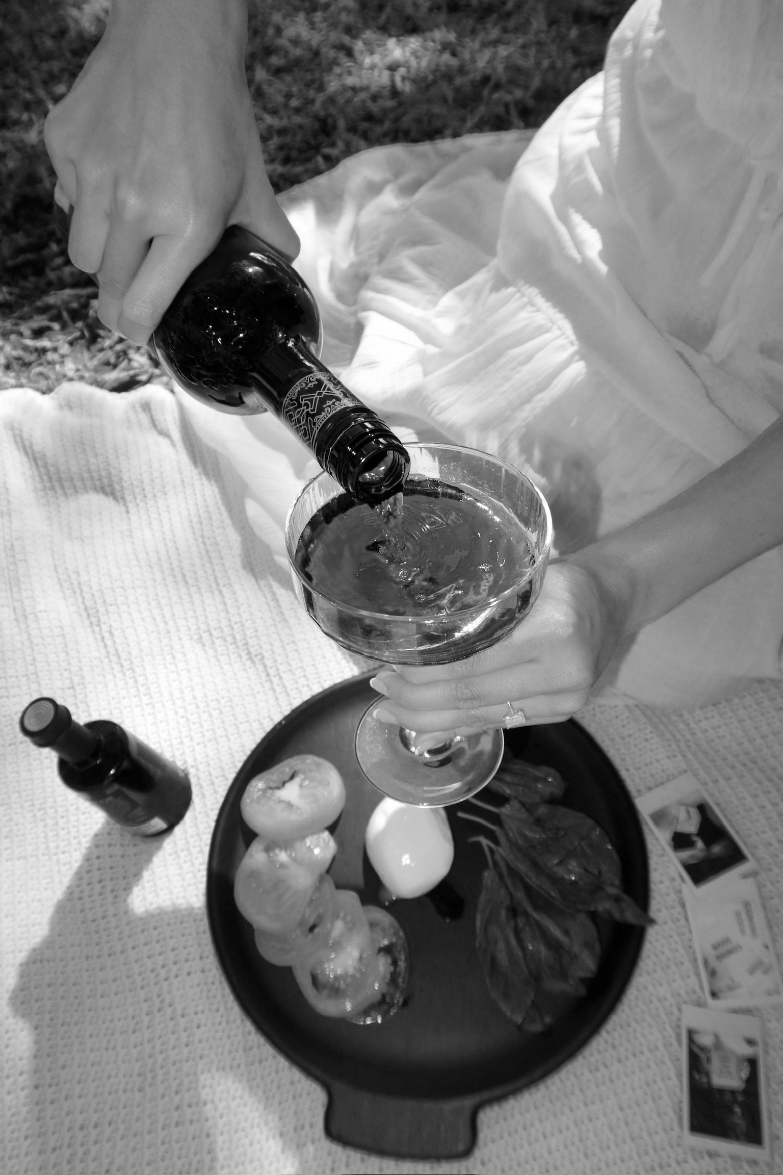 A person pours a beverage into a wine glass during a picnic, with a tray of food including tomatoes, basil, and what appears to be mozzarella cheese, on a tray nearby.