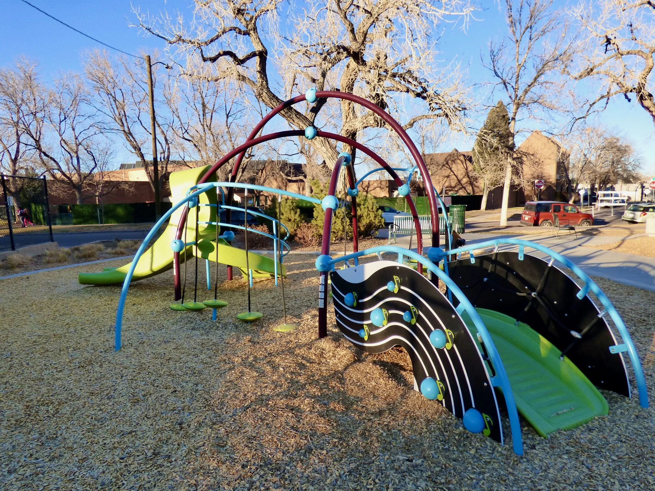 Colorful playground with slides and climbing structures in a park during winter with leafless trees and parked cars in the background.