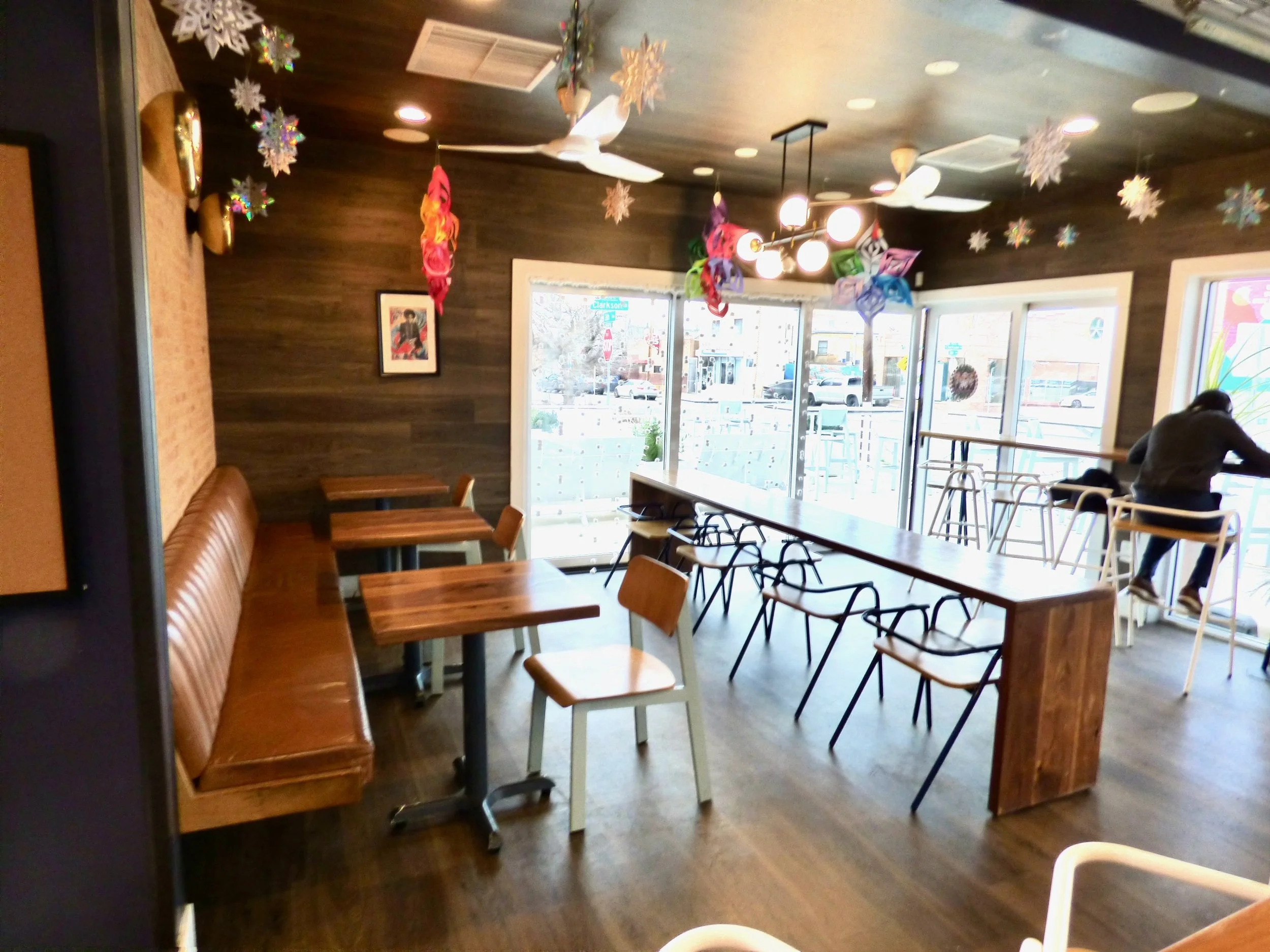 Empty indoor café with wooden tables and chairs, decorated for winter with snowflakes and colorful hanging ornaments hanging from the ceiling, and large windows letting in natural light.