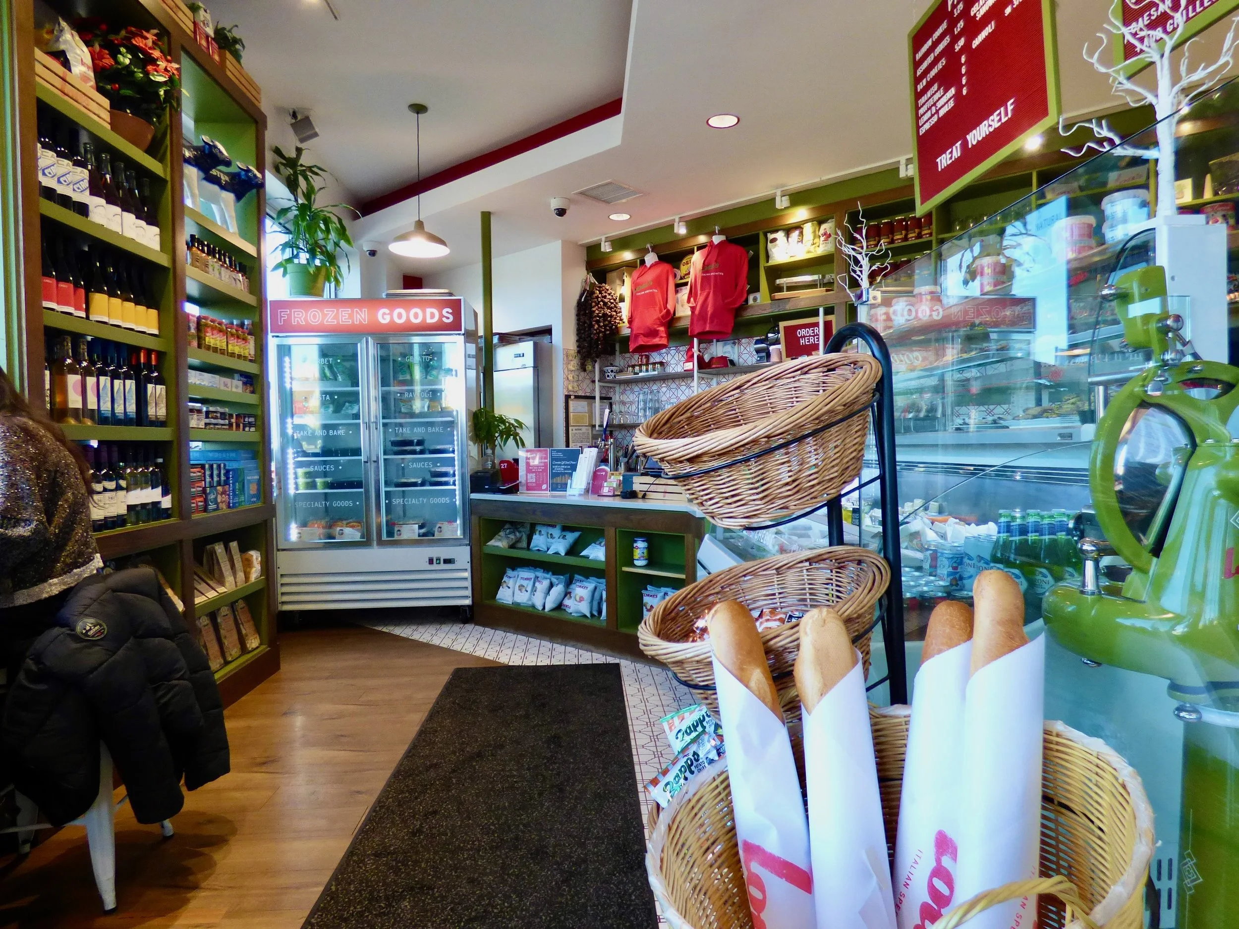 Interior of a small grocery or deli with shelves stocked with bottles, jars, and packaged goods, a refrigerated section labeled "Frozen Goods," baskets with baguettes, a green coffee machine, and a display case with refrigerated products.