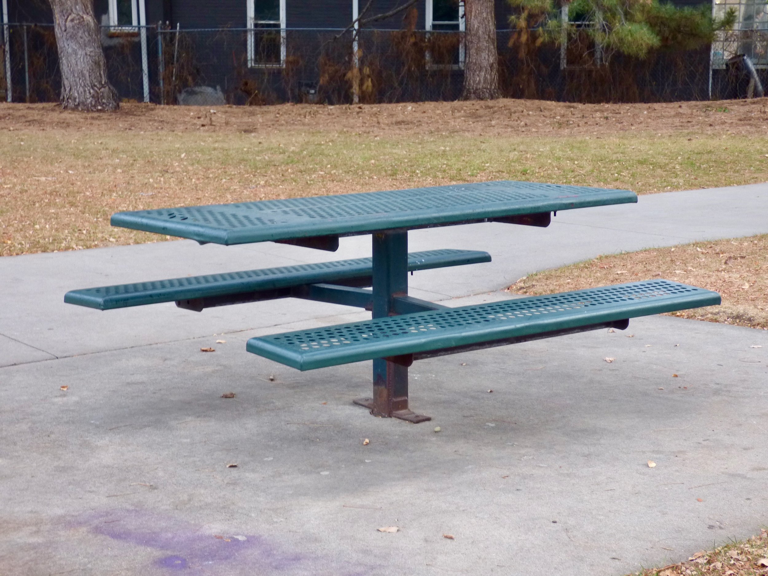 A green metal picnic table with attached benches on a concrete sidewalk in a park, with grass, trees, and a chain-link fence in the background at Frederick Douglas Park, Denver, CO 80205.