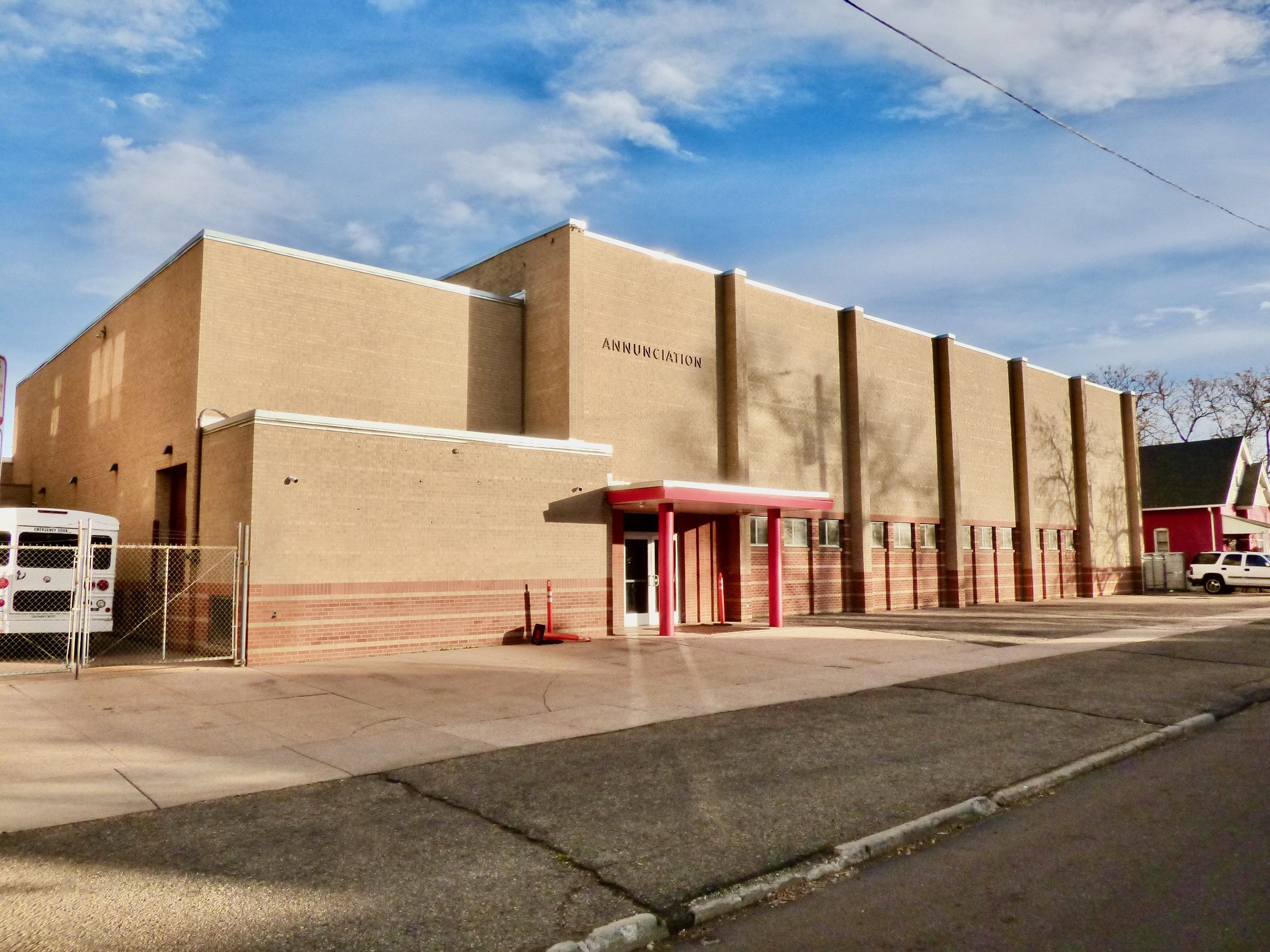 Exterior of a beige brick building with the word "ANNUNCIATION" on the wall, featuring a red entrance awning, located on a street with parked vehicles and a chain-link fence on the left.