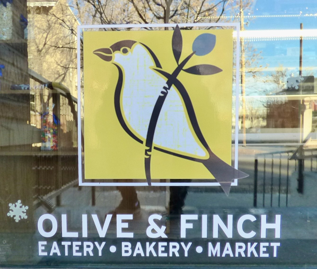Sign for Olive & Finch, an eatery, bakery, and market, featuring a graphic of a finch with an olive branch on a yellow background