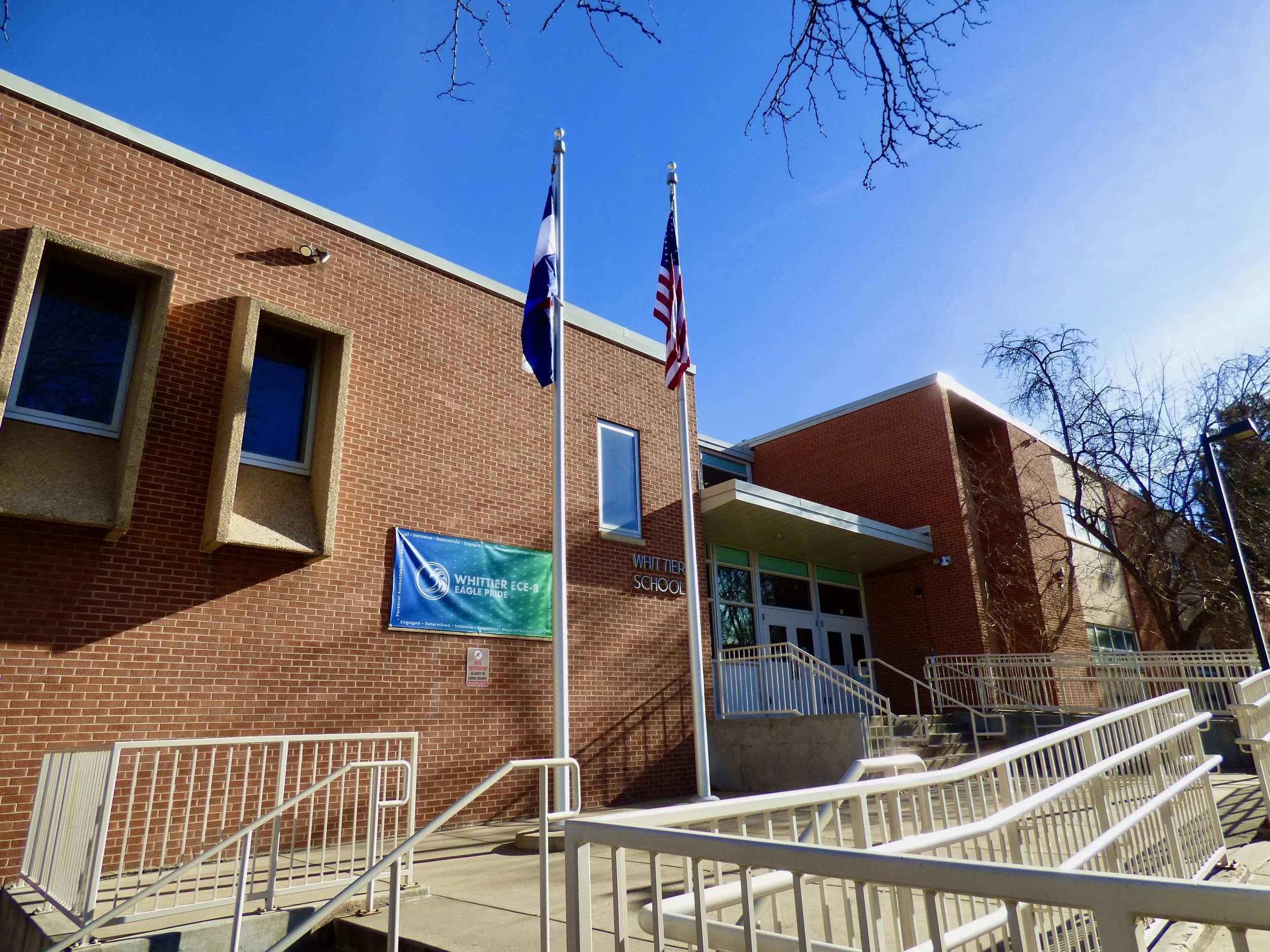 Exterior of Whittier School with brick walls, flagpoles with American and another flag, and a blue sign that reads 'Whittier ECE-8 Eagle Pride.' Ramps lead to the entrance, and leafless trees at Whittier Elementary School, Denver, CO 80205.