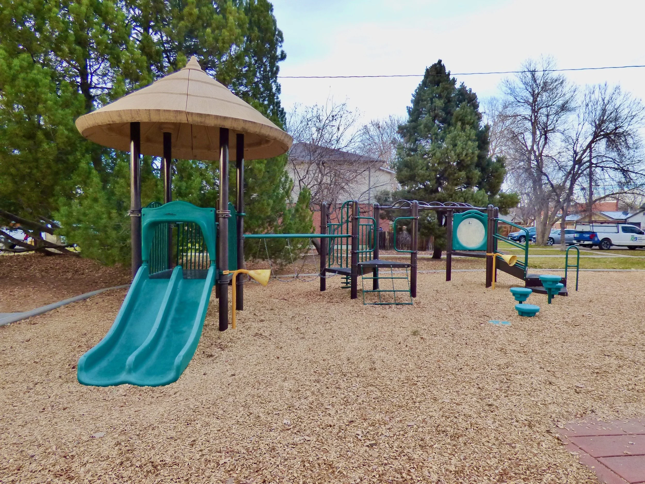 Empty playground with a slide, climbing equipment, and springy seats, surrounded by trees and houses in the background at Frederick Douglas Park, Denver, CO 80205.