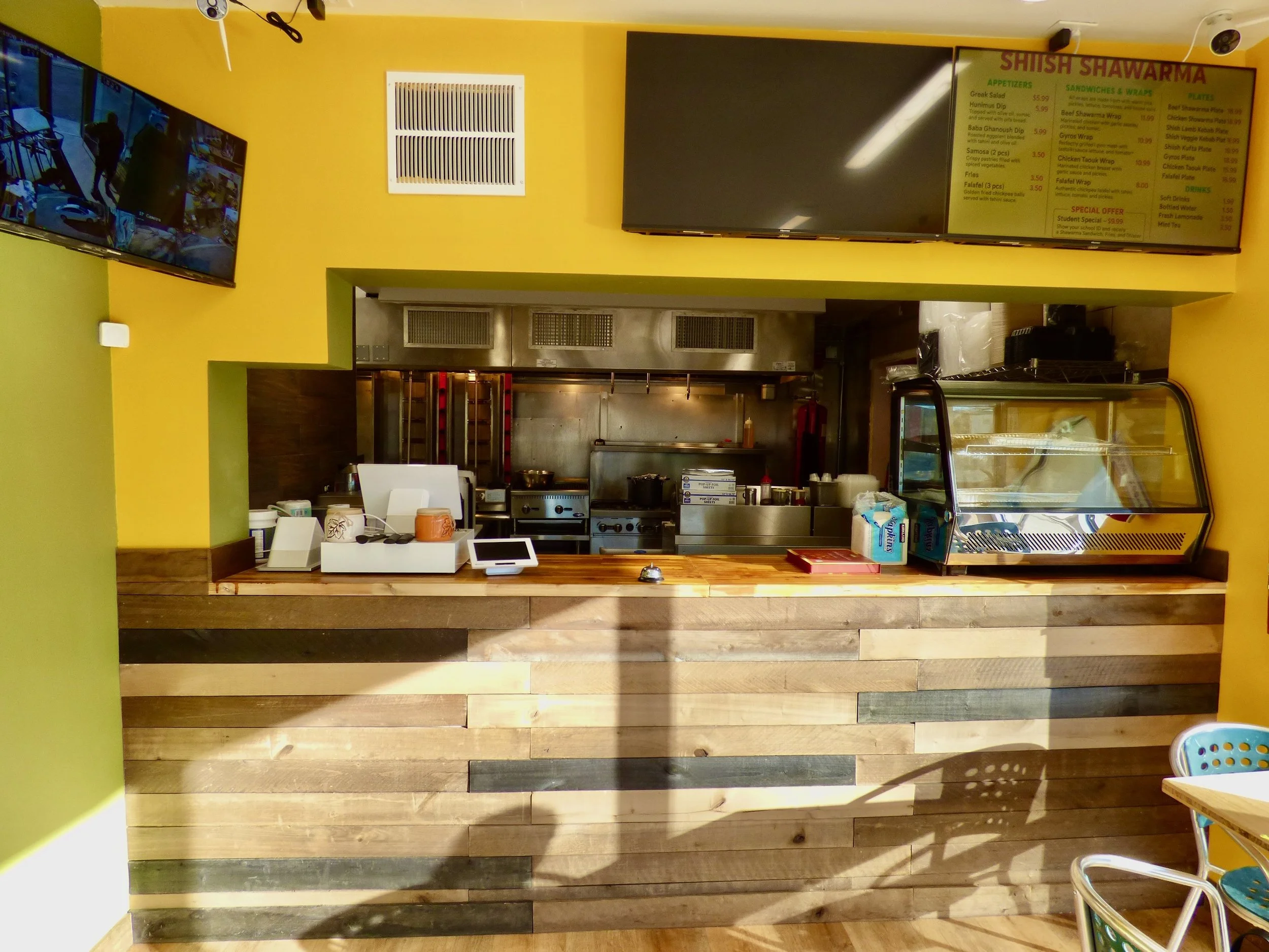 The image shows a restaurant counter with a bright yellow wall behind it. There is a large menu board hanging above the counter, and a flat-screen TV is mounted next to it, displaying multiple camera feeds. The kitchen area is visible behind the counter, with stainless steel appliances and tools. The counter itself is made of wooden planks, and there are some small items like a cash register, tissue box, and trays on it. To the right, there is a glass display case, and a portion of a colorful chair is visible in the bottom right corner.