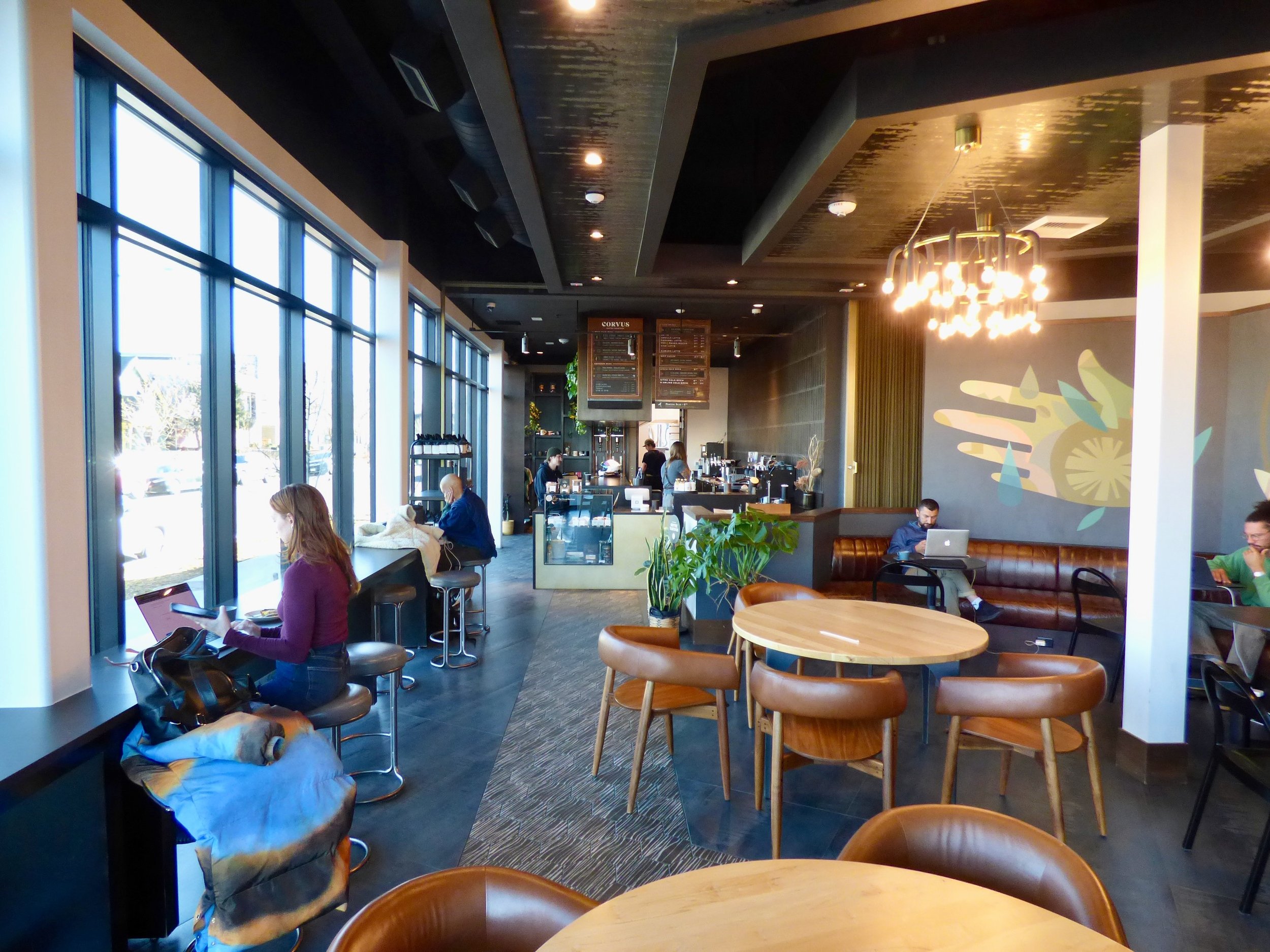 Interior of a modern coffee shop with large windows, wooden tables and chairs, customers working on laptops, and a barista counter in the background at Elemental Bakery & Coffeehouse, Denver, CO 80205.