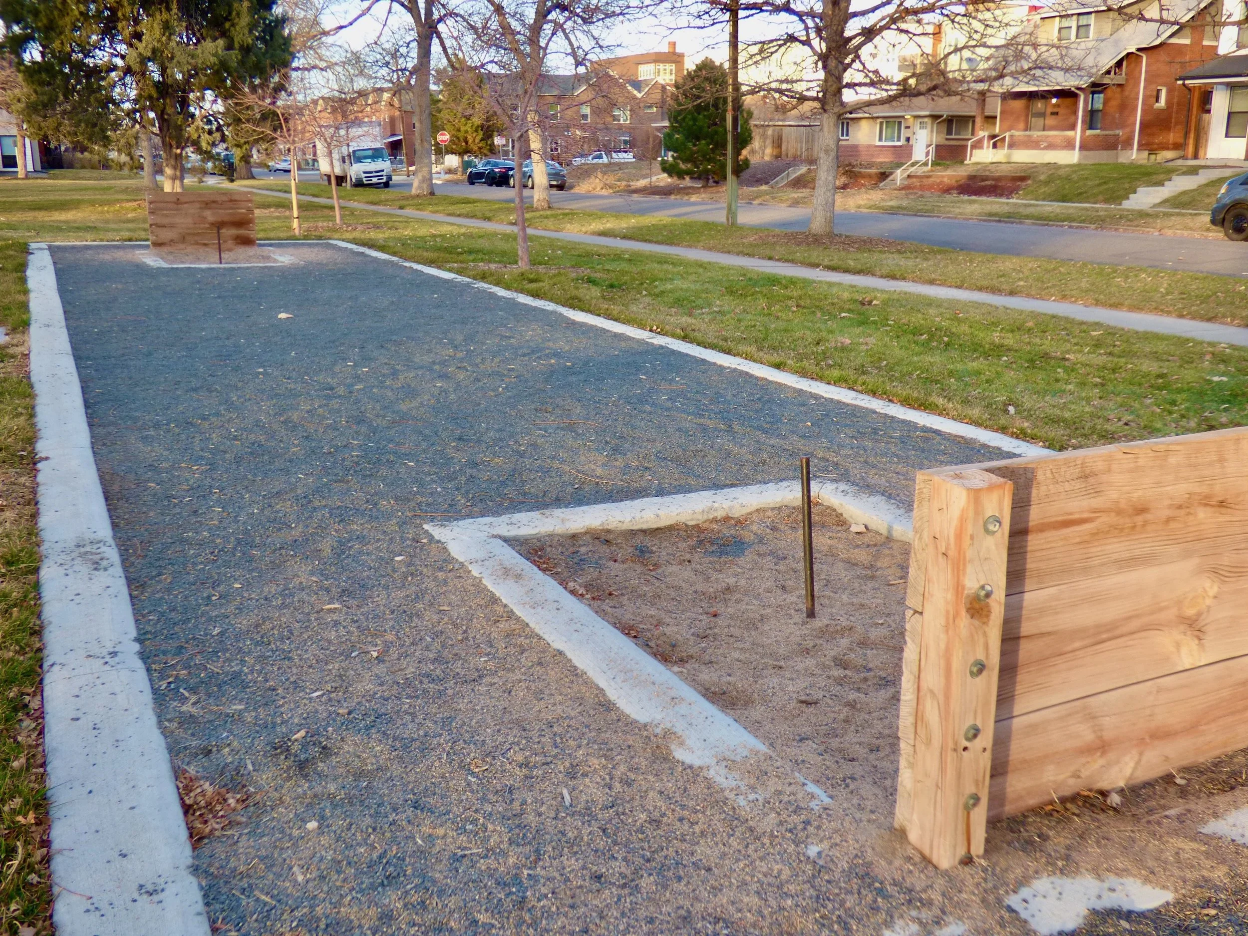 A horseshoe pit with a sand trap and a raised wooden barrier at the edge, located outdoors in a neighborhood park with trees, grass, and houses in the background in Russell Square Park, Denver, CO 80205.