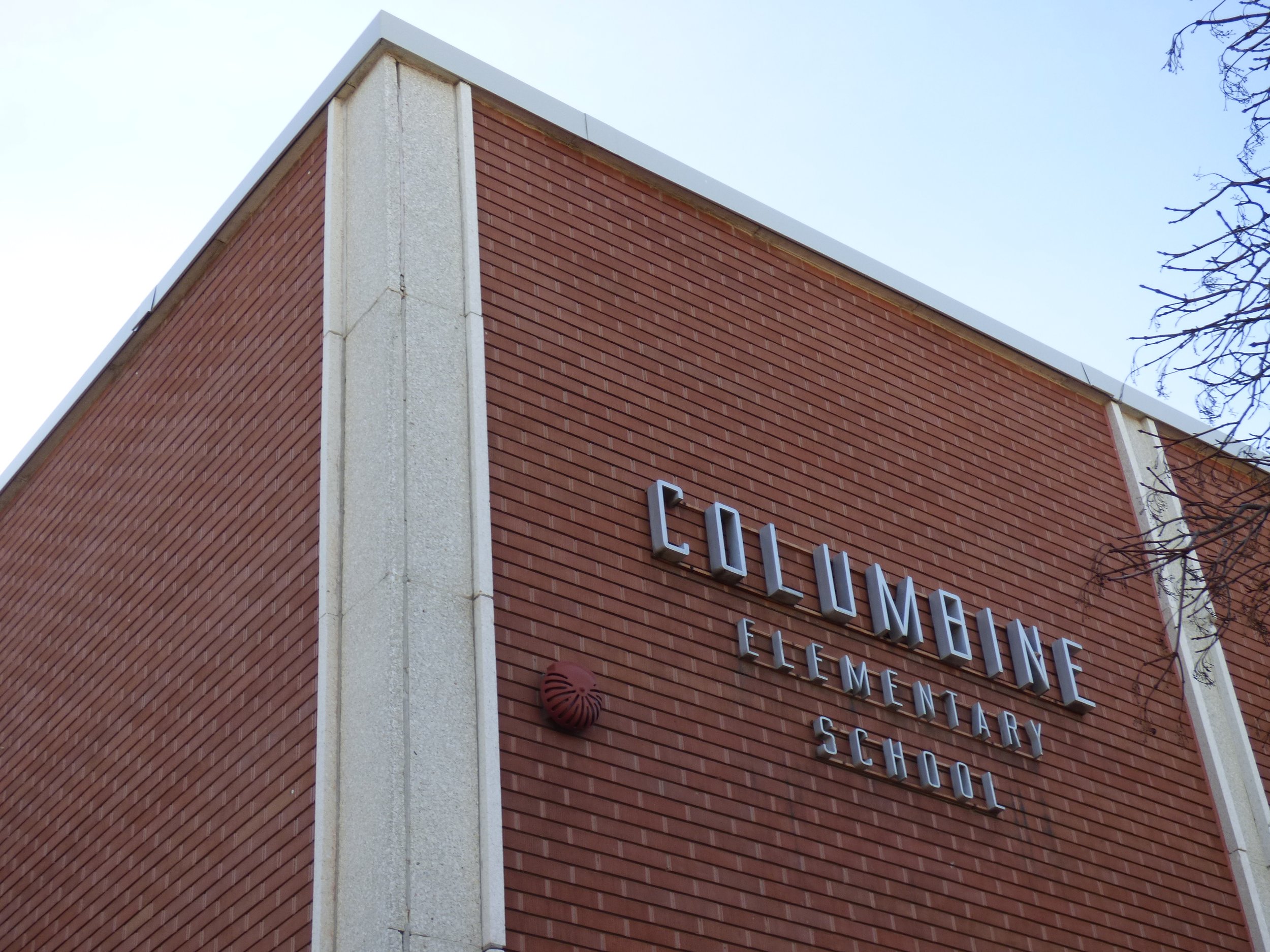 Exterior of Columbine Elementary School with brick facade, metal letter signage, and a corner of the building visible against a clear sky.