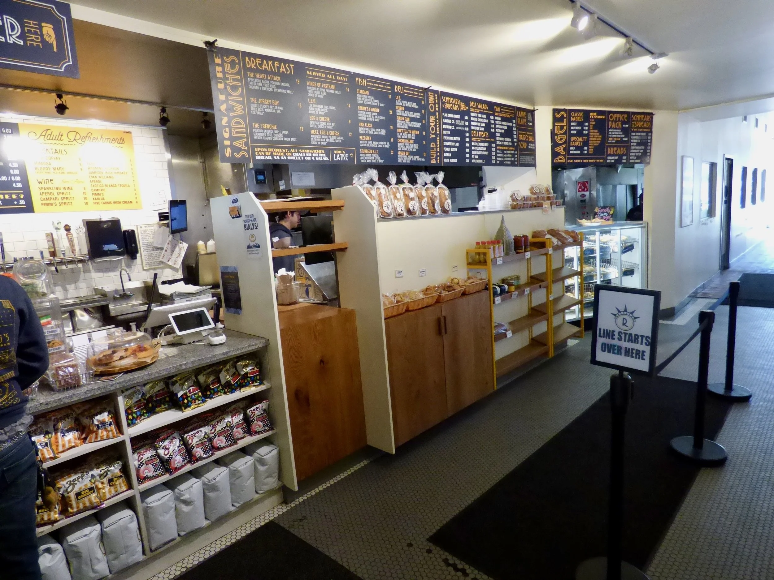 Inside a deli or cafe, showing a counter with baked goods, shelves with snacks, and a menu board above. A sign indicates the line starts here.