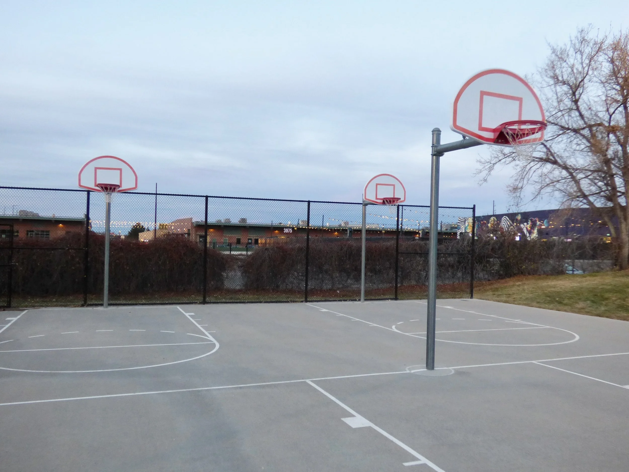 Empty outdoor basketball court with three hoops, surrounded by a black fence and a tree, under a cloudy sky during dusk in Martin J. Schafer Park, Denver, CO 80205.