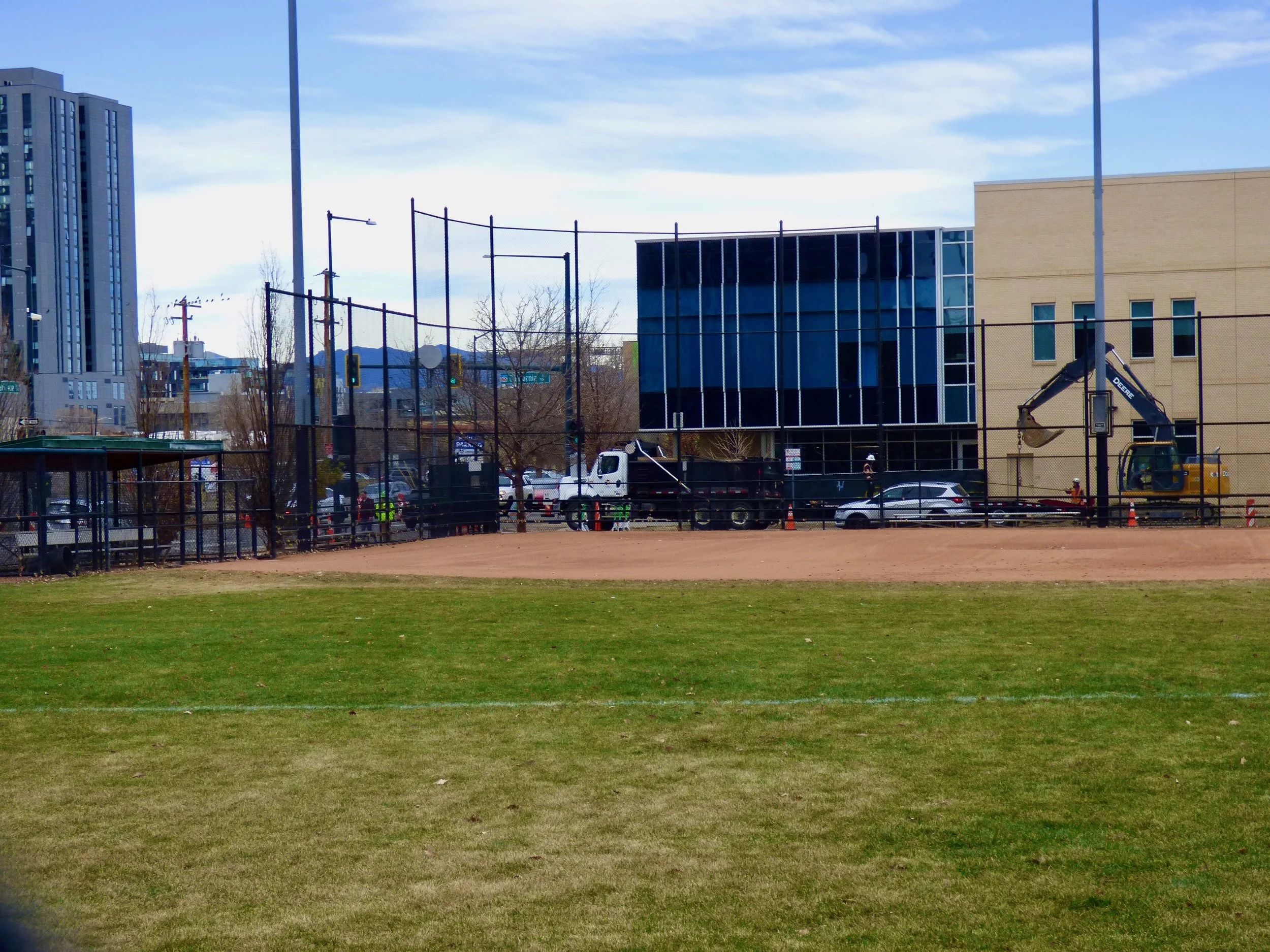 A baseball field in the foreground with a construction site beyond the fence, including machinery and buildings, and a cityscape in the background in Sonny Lawson Park, Denver, CO 80205.