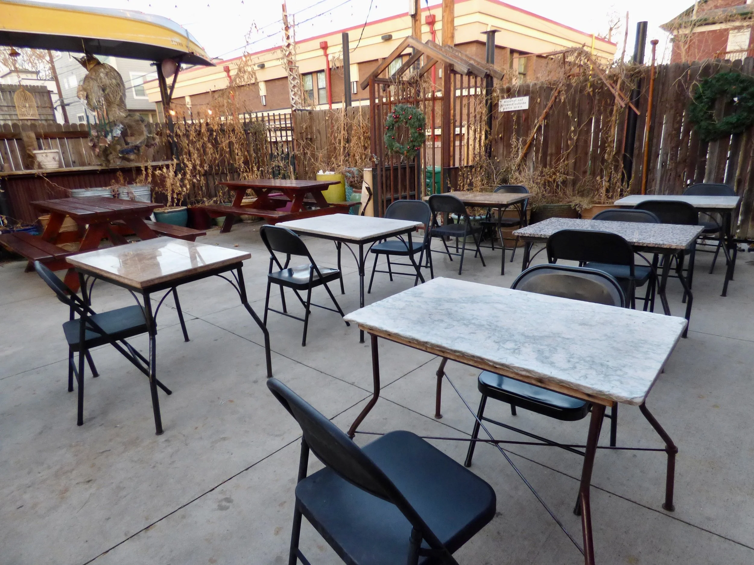 Empty outdoor patio with multiple tables and chairs, some with granite or marble tops, enclosed by a wooden fence adorned with holiday wreaths and dried plants at The Thin Man, Denver, CO.
