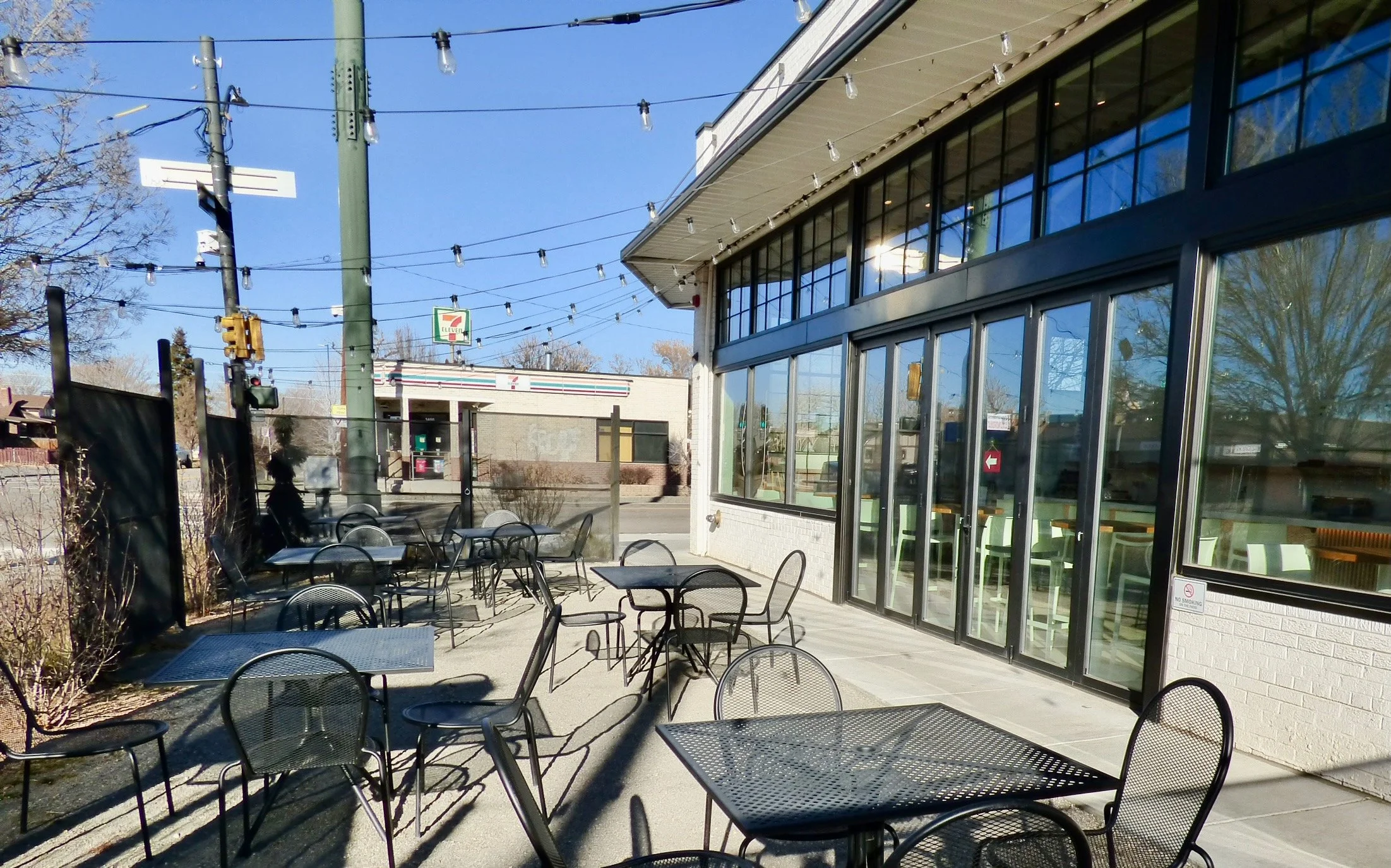 Outdoor seating area with black metal tables and chairs outside a restaurant or café, with a large glass window and door, and a 7-Eleven in the background under a clear blue sky at Hot Shot Coffee Shop, Denver, CO 80205.