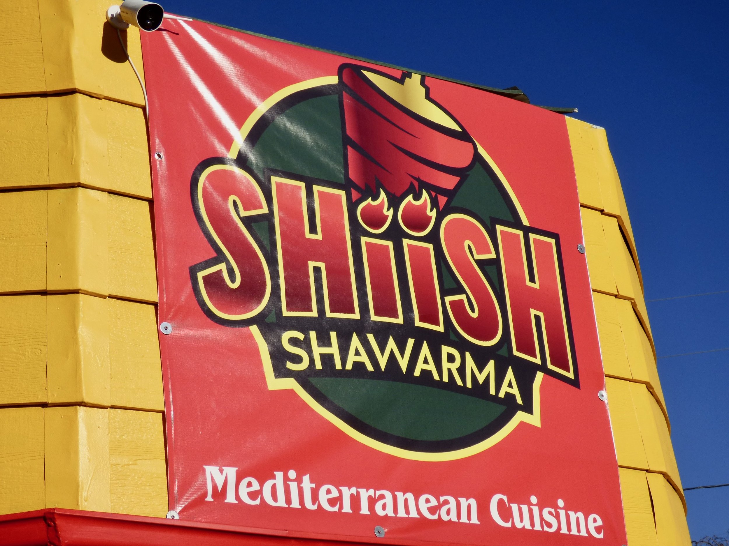 A red sign with black, yellow, and white text for Shishi Shawarma on a yellow wall, advertising Mediterranean cuisine. The sign features a logo with a shawarma wrap and flames, and a surveillance camera is mounted nearby.