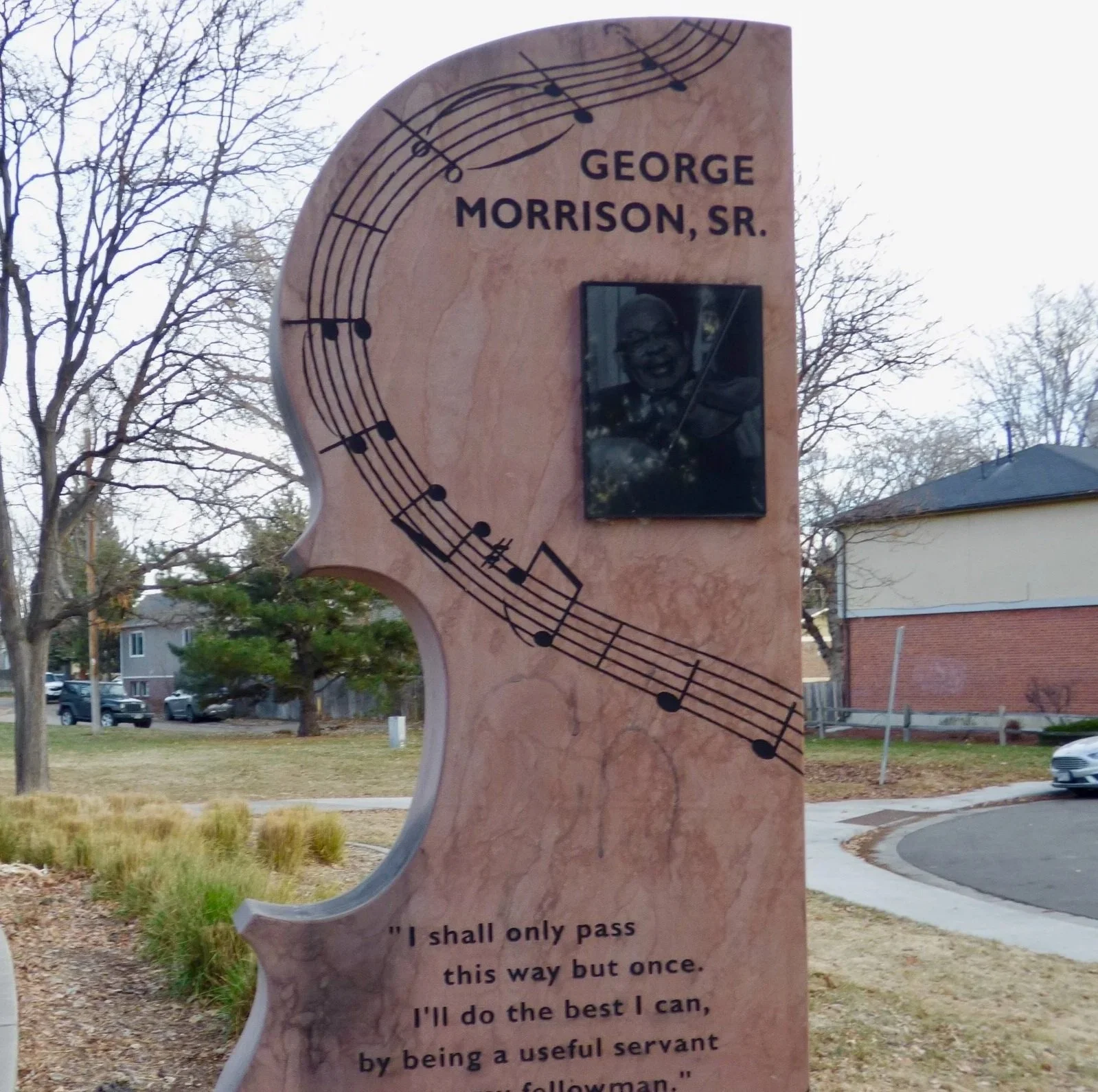 A memorial monument in the shape of a violin with musical notes printed on it. It features a black-and-white photo of a smiling man playing the violin and an inscription that reads, "I shall only pass this way but once. I'll do the best I can, by being a useful servant and follower." The background includes trees, houses, and parked cars in a suburban neighborhood.
