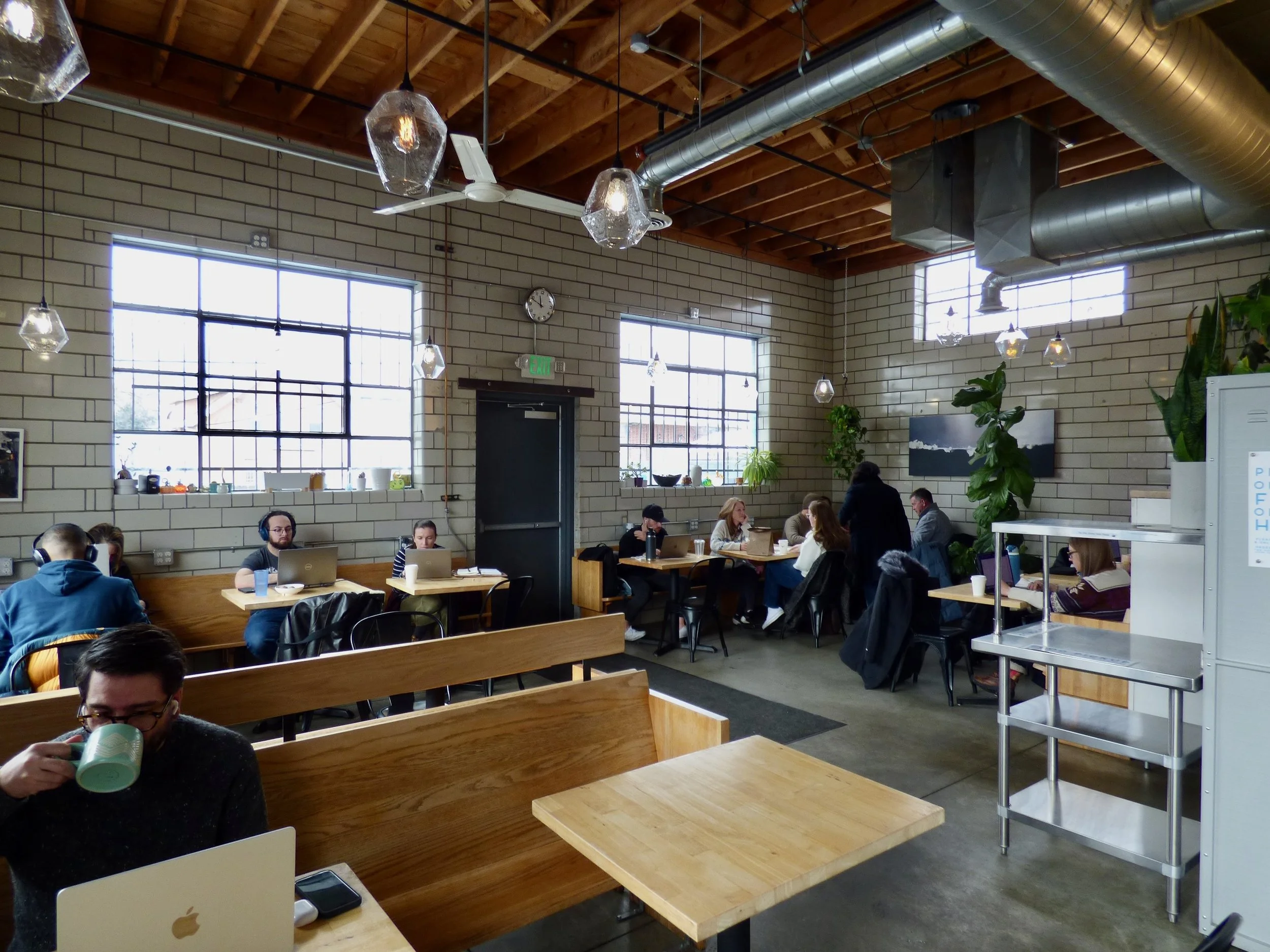 People working in a modern café with large windows, exposed brick walls, and wooden ceiling beams, some using laptops, others drinking coffee.
