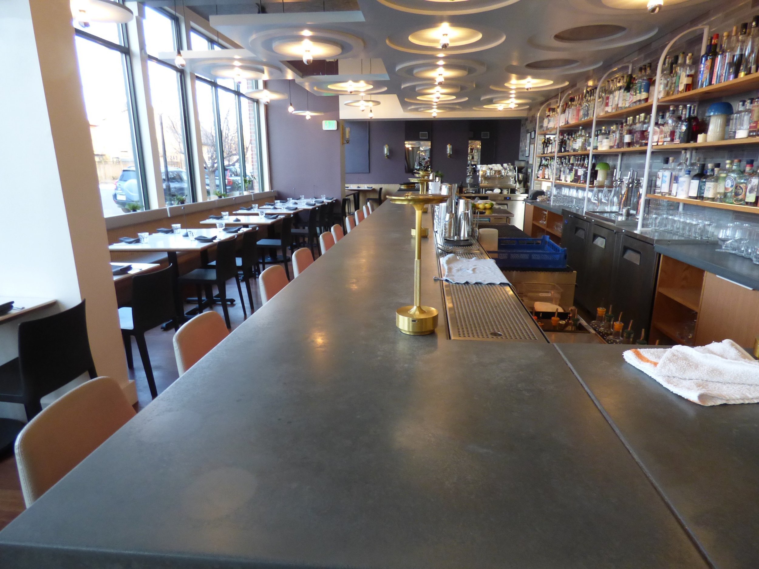 Empty restaurant with a long bar counter, tables set with napkins and glasses, large windows, and a well-stocked bar with bottles behind it at Point Easy, Denver, CO 80205.