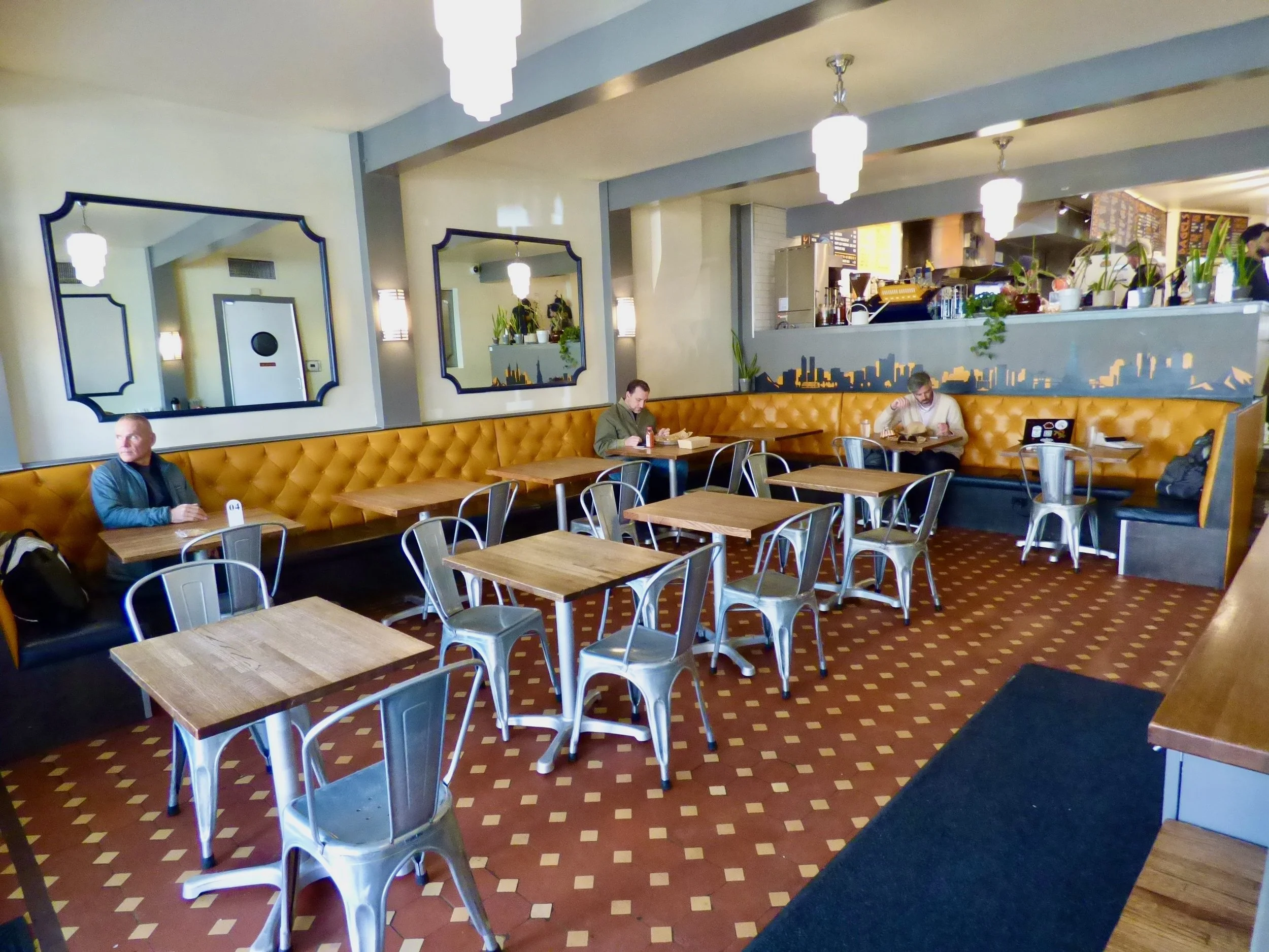 Interior of a cozy coffee shop with wooden tables, metal chairs, a long mustard-colored booth with tufted upholstery, and three customers seated and reading or working. There are large mirrors on the wall, hanging pendant lights, and a small cityscape mural behind the booth.