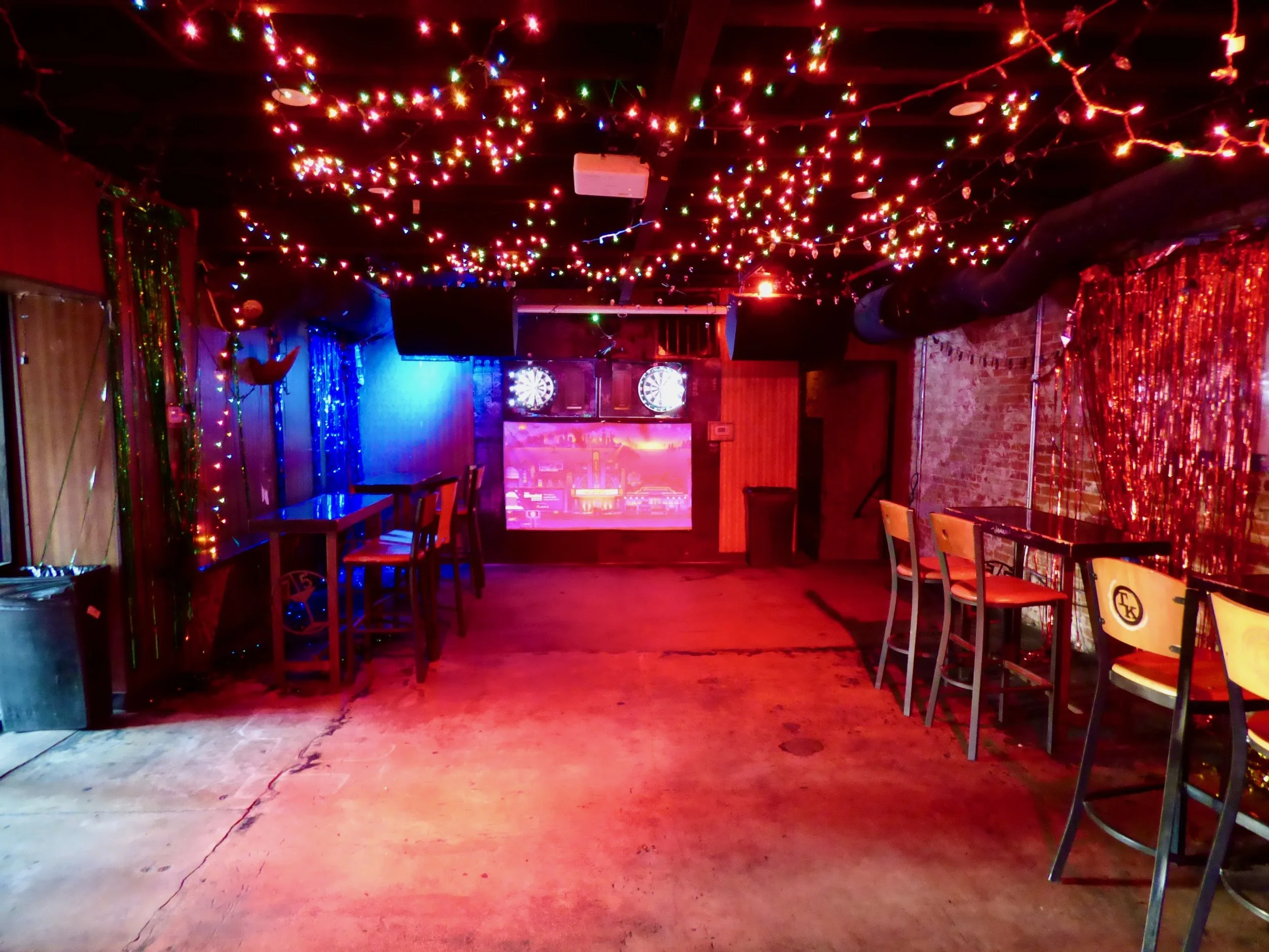 Empty entertainment venue decorated with colorful string lights on the ceiling, fairy lights on the walls, a mural on the back wall, and dartboards on the wall at 715 Club, Denver, CO 80205.