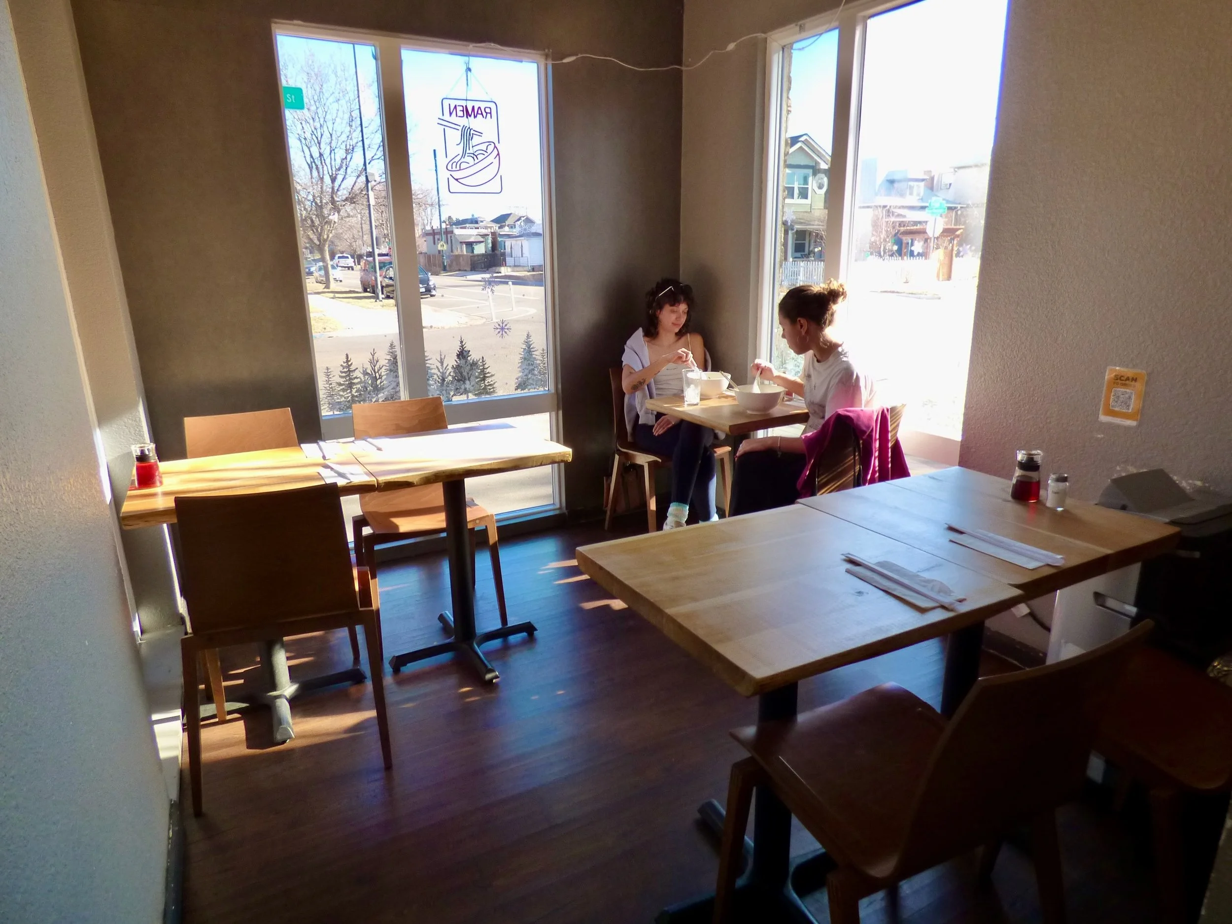 Two women sitting at a small table in a sunlit cafe, eating and talking. Large windows show a street view with trees and houses outside.