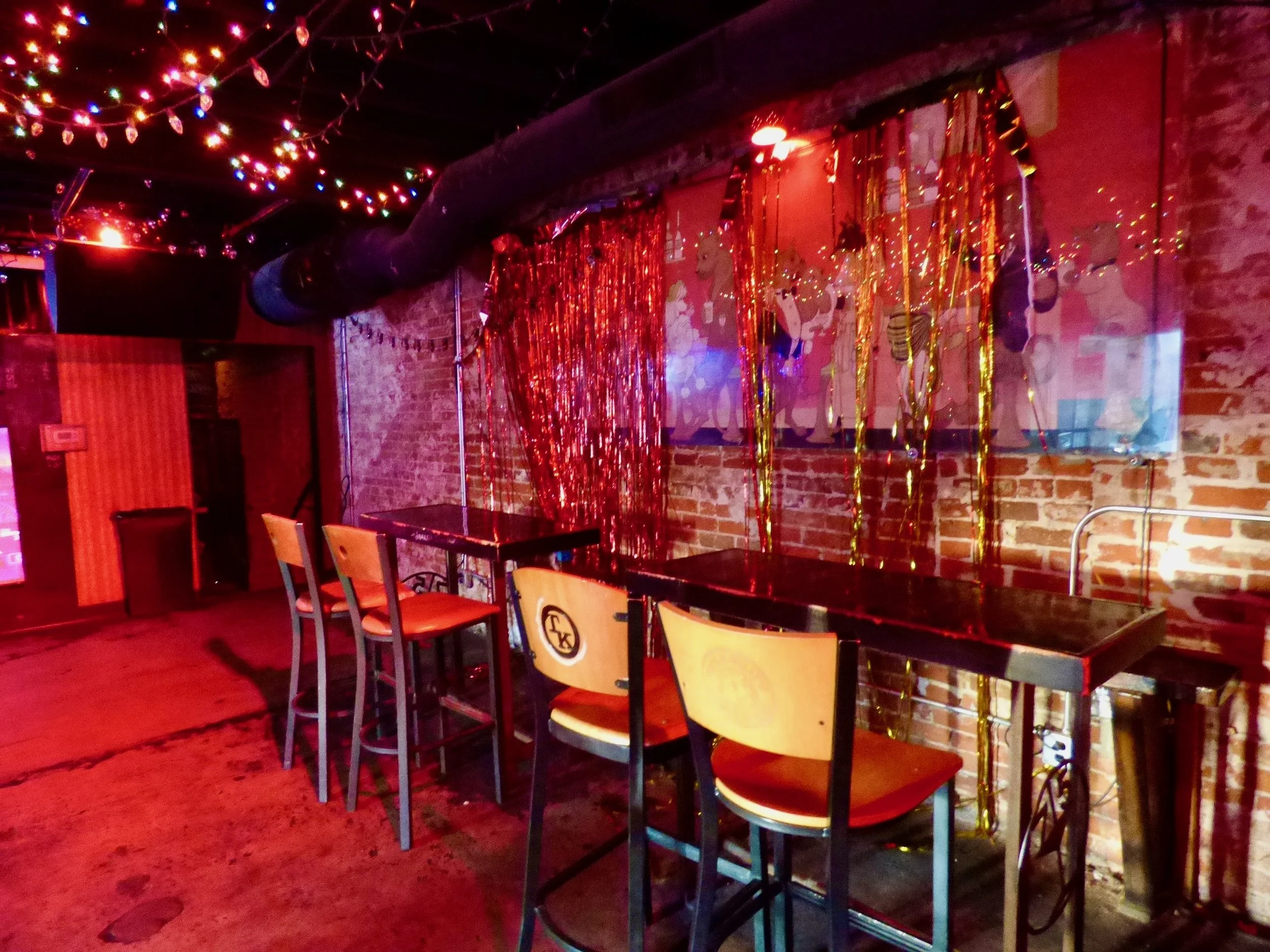 Bar area decorated with colorful string lights, tinsel streamers, and a mural of playful cartoon bears on an exposed brick wall, with cocktail tables and chairs at 715 Club, Denver, CO 80205.