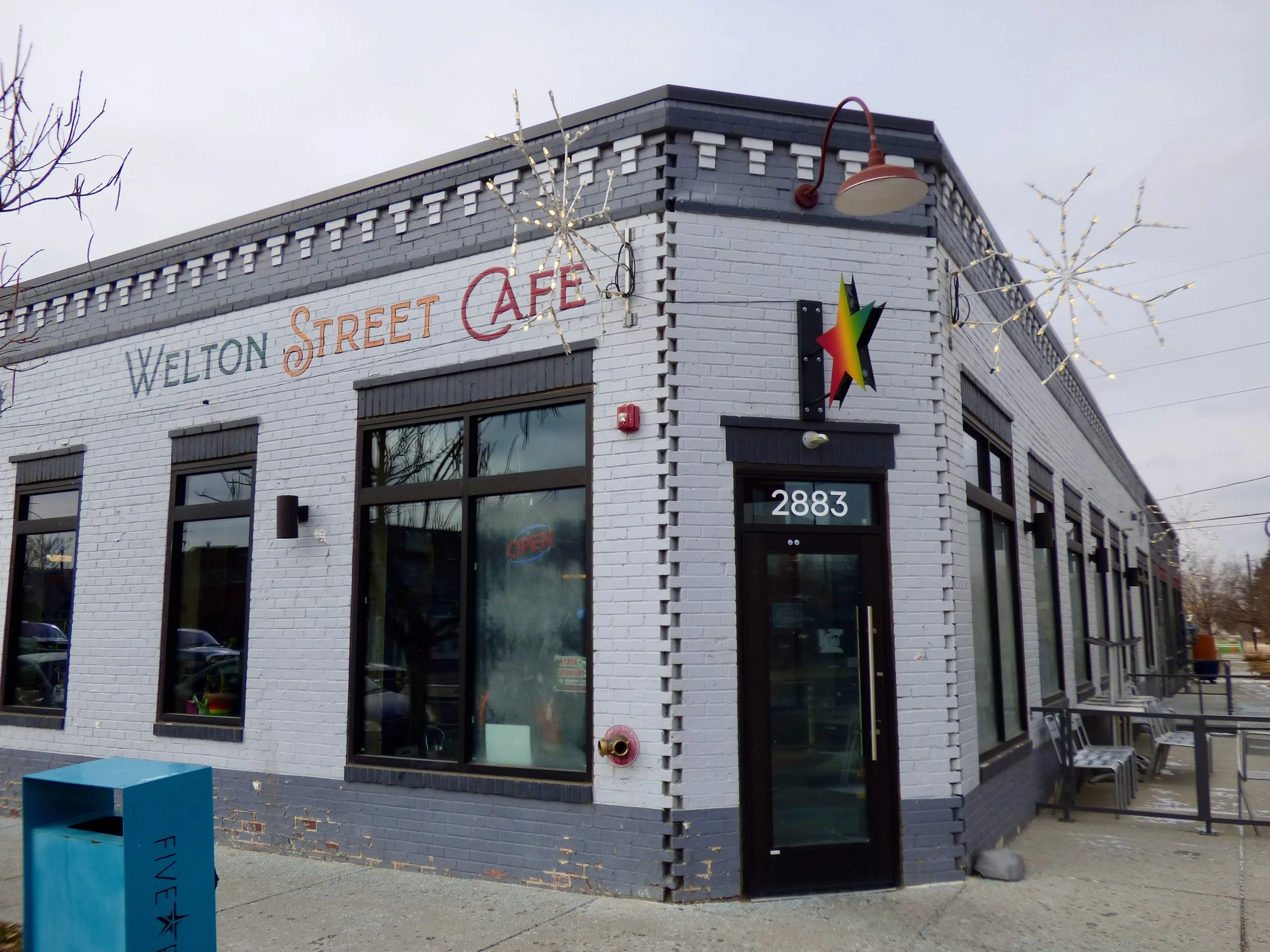 White building with black trim, pink and orange sign reading "Welton Street Cafe," large windows, outdoor seating, and holiday lights on the roof, located at 2883.