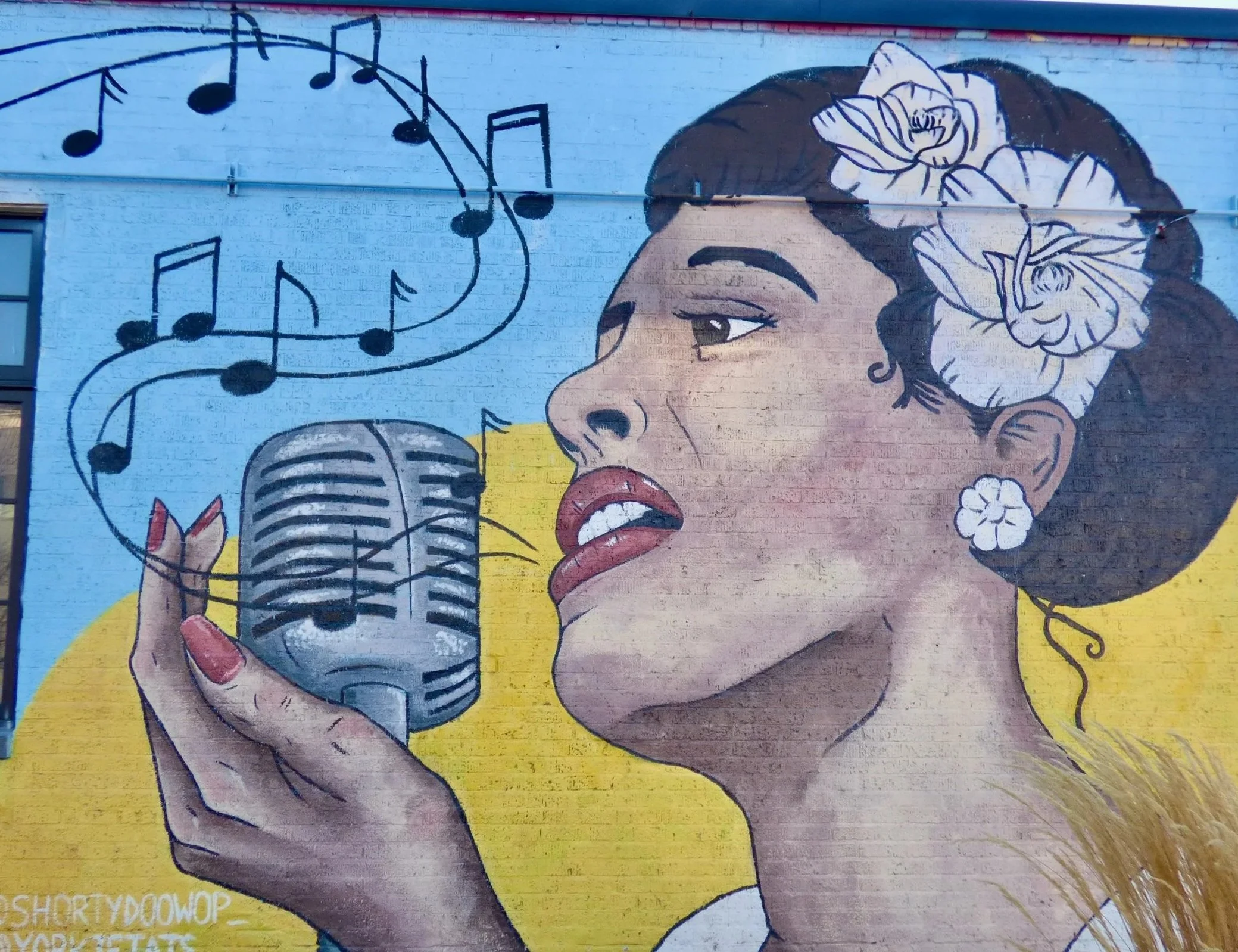 Mural of a woman singing into a vintage microphone, with musical notes floating above, grass earrings, flowers in her hair, and a yellow background.