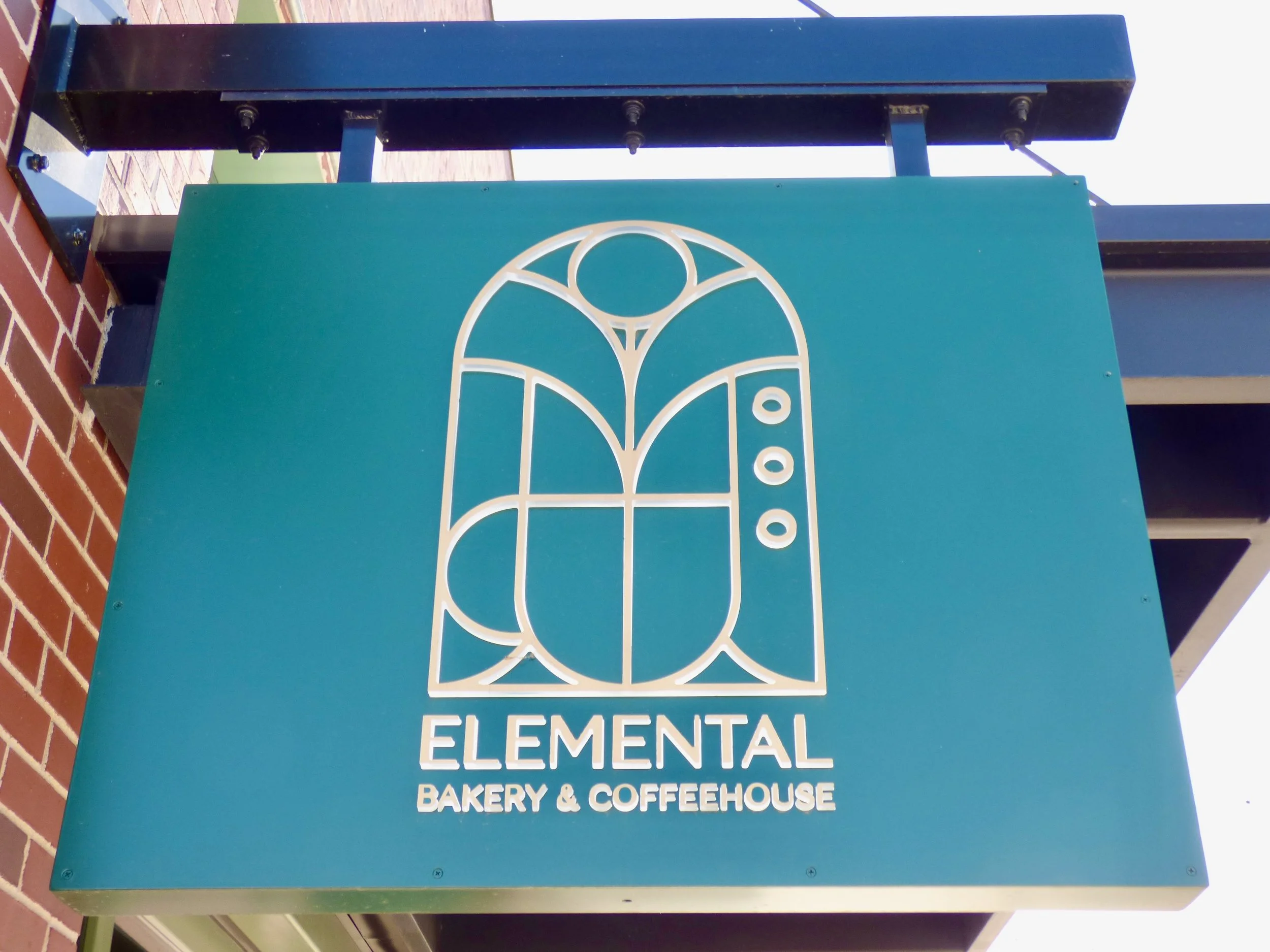 A teal business sign for Elemental Bakery & Coffeehouse with a minimalist white line art logo featuring stylized shapes and the text below at Elemental Bakery & Coffeehouse, Denver, CO 80205.