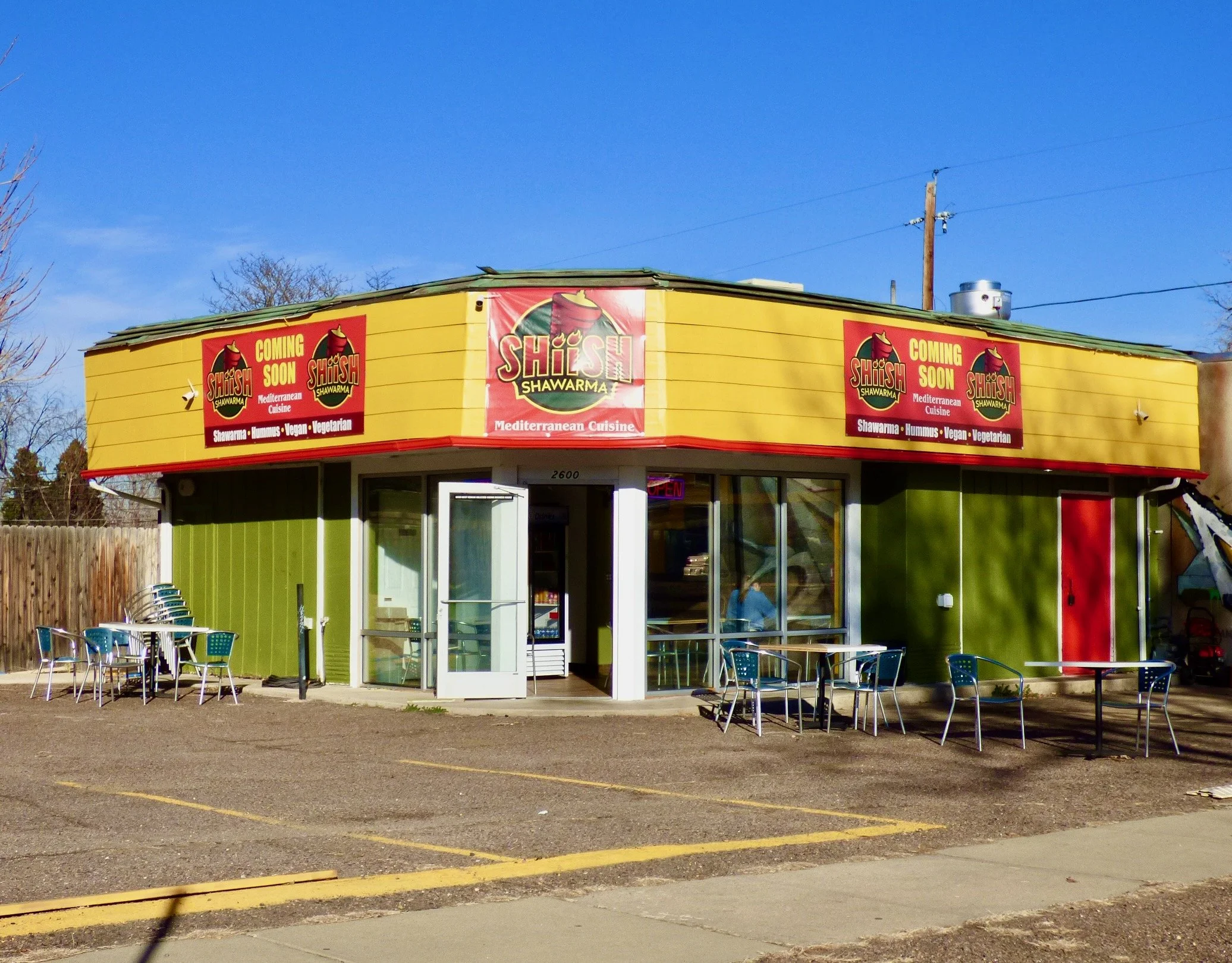 Colorful building with outdoor seating, advertising Mediterranean cuisine, shawarma, hummus, vegan, and vegetarian options, with signs indicating opening soon.