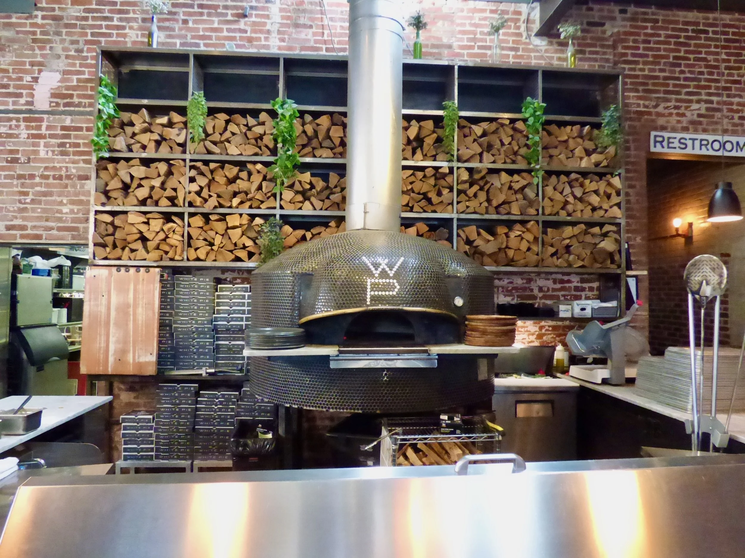 Wood-fired pizza oven in a restaurant with stacked wood storage and brick walls.
