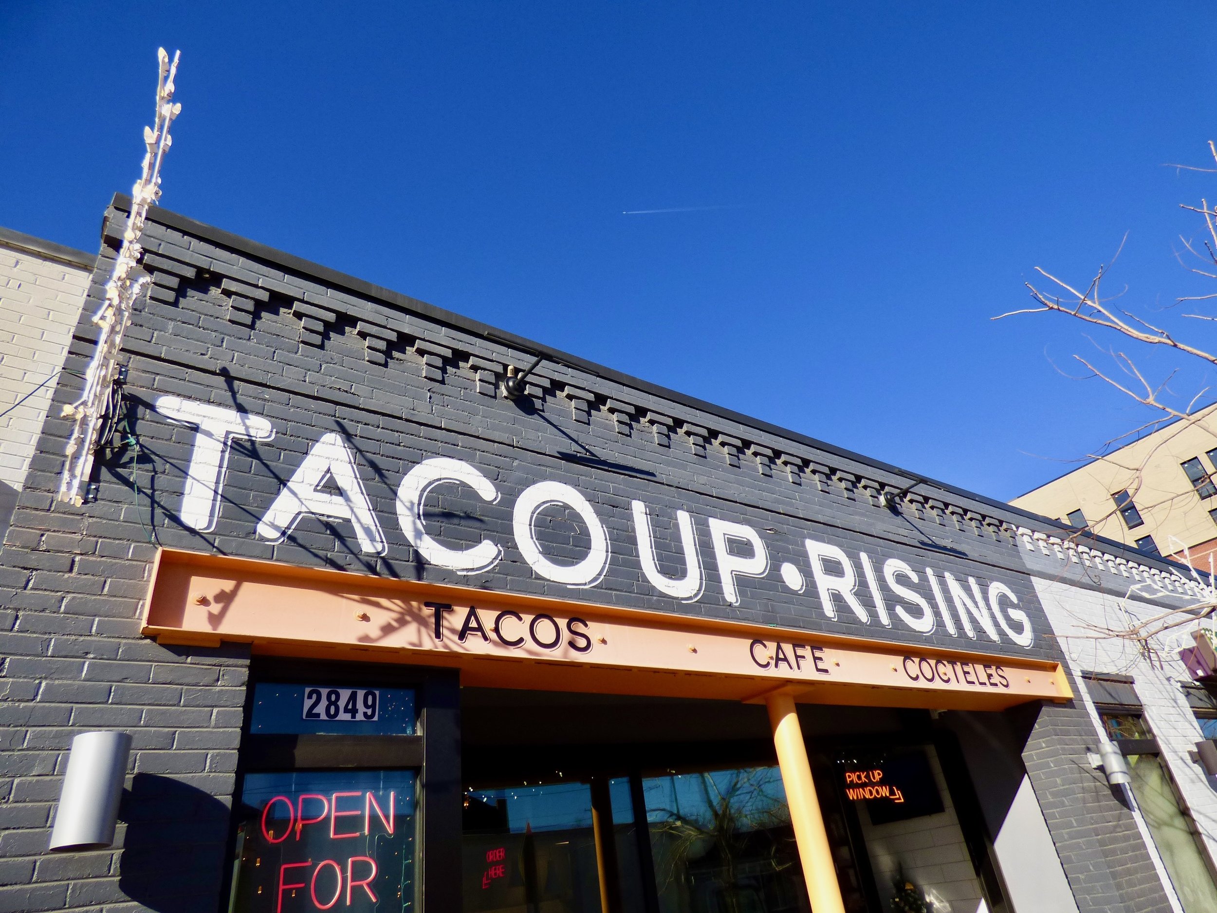 Exterior of a restaurant named 'TACUP RISING' with a sign advertising tacos, cafe, and cocktails. The building has a black painted brick facade with a bright orange awning and signs that say 'Open for'. The sky is blue with clear weather.
