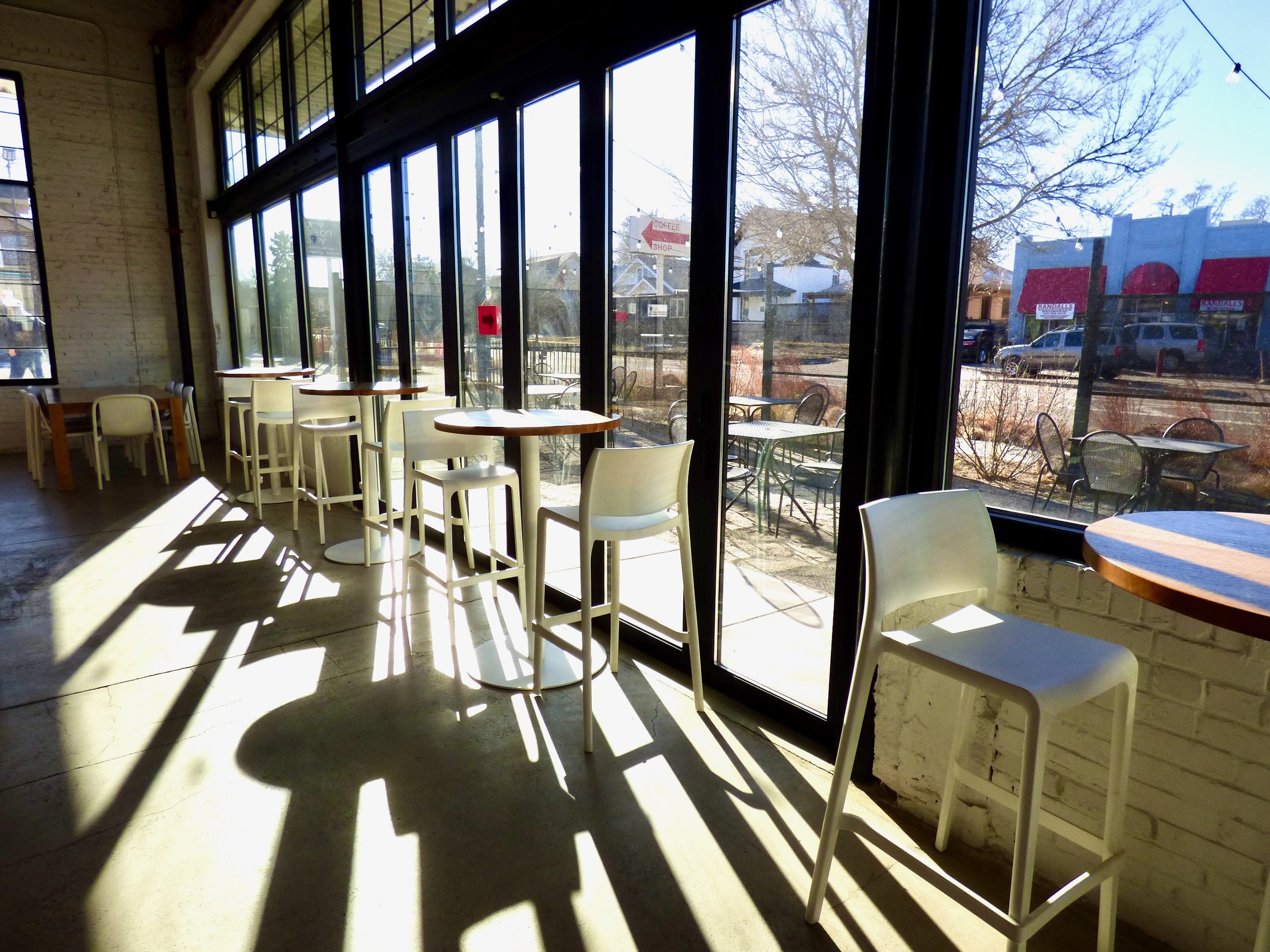 Empty cafe with high tables and chairs near large windows, sunlight casting shadows inside, outdoor seating visible through the windows at Hot Shot Coffee Shop, Denver, CO 80205.