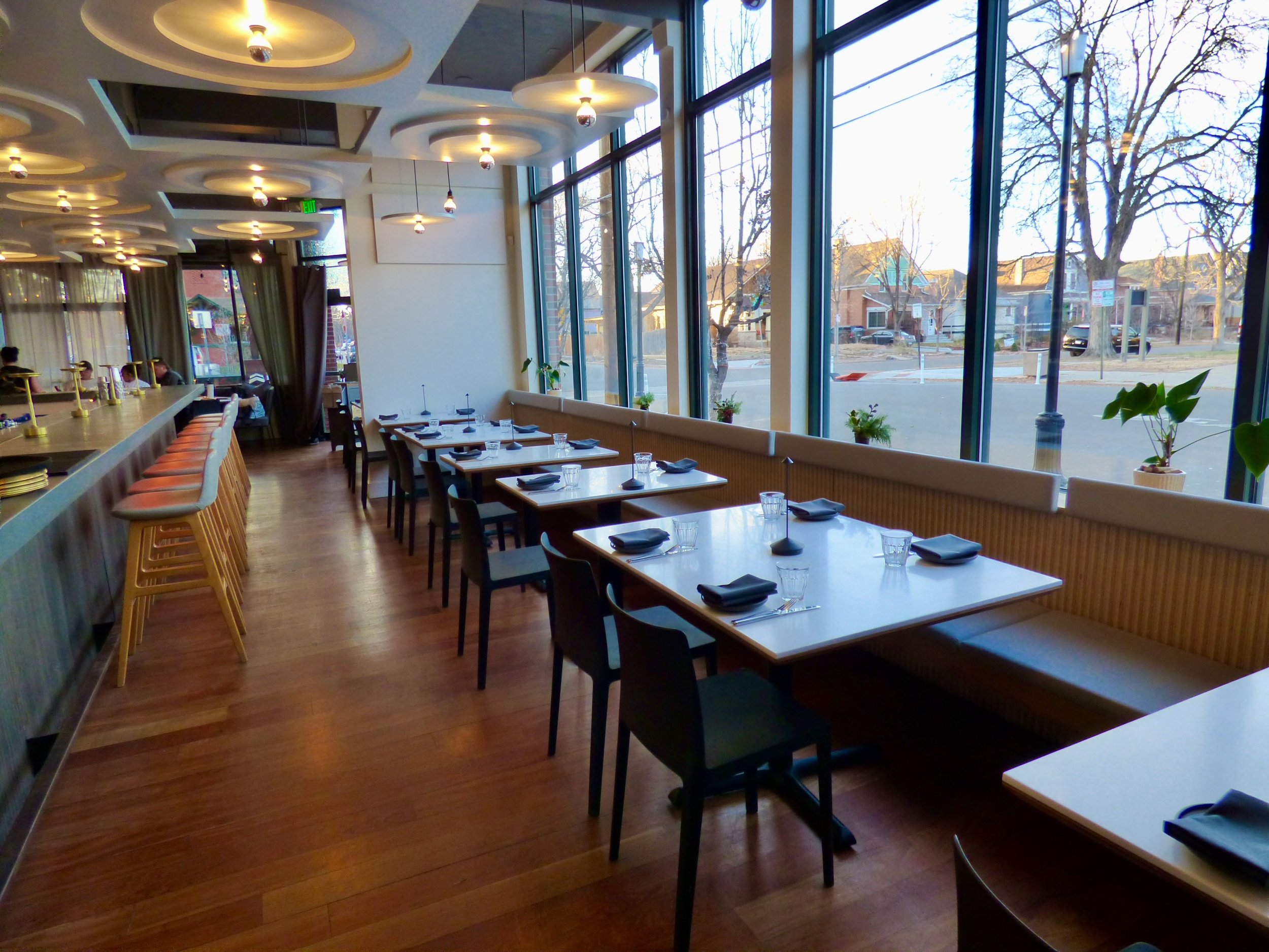 An empty restaurant dining area with large floor-to-ceiling windows showing an outdoor street view, set with black napkins, glasses, and cutlery on the tables, and wooden flooring at Point Easy, Denver, CO 80205.