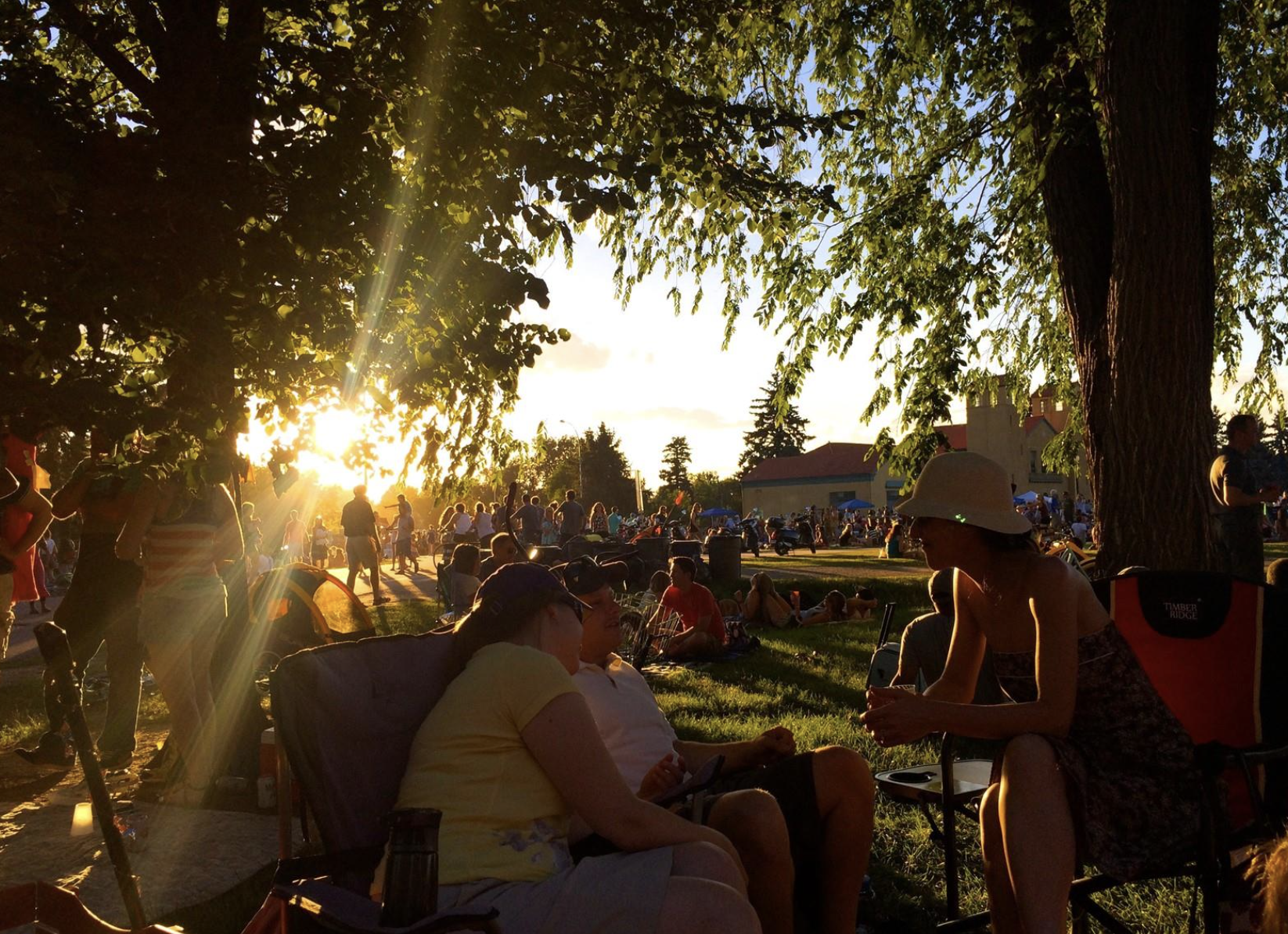 People gathered outdoors in a park during sunset. Some are sitting, some walking, and others socializing under trees during Jazz in the Park at City Park, Denver, CO.