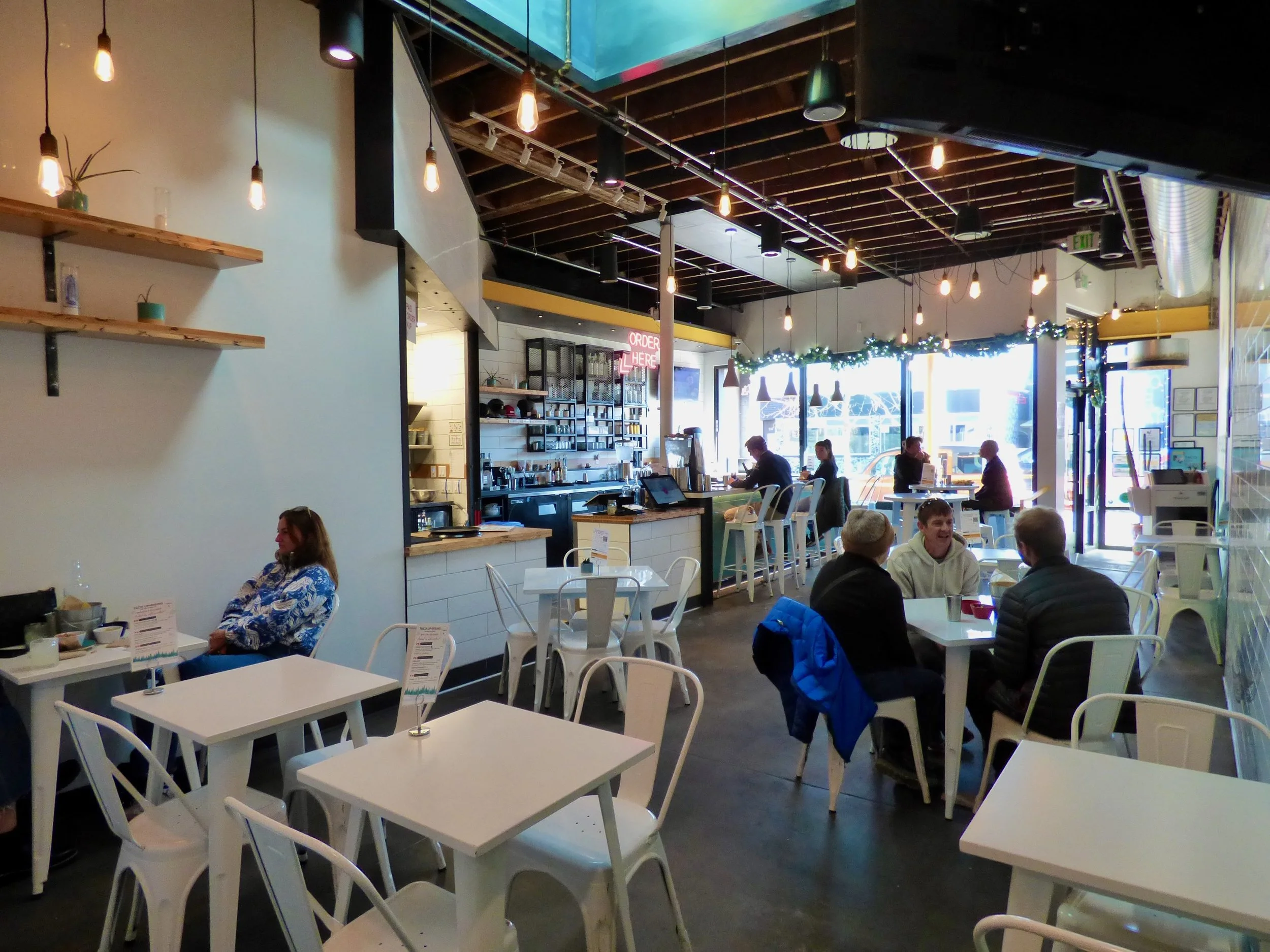 Interior of a modern cafe with white tables and chairs, some people sitting and talking, large windows at the back with decorated garlands, pendant lights hanging from exposed wooden beams.