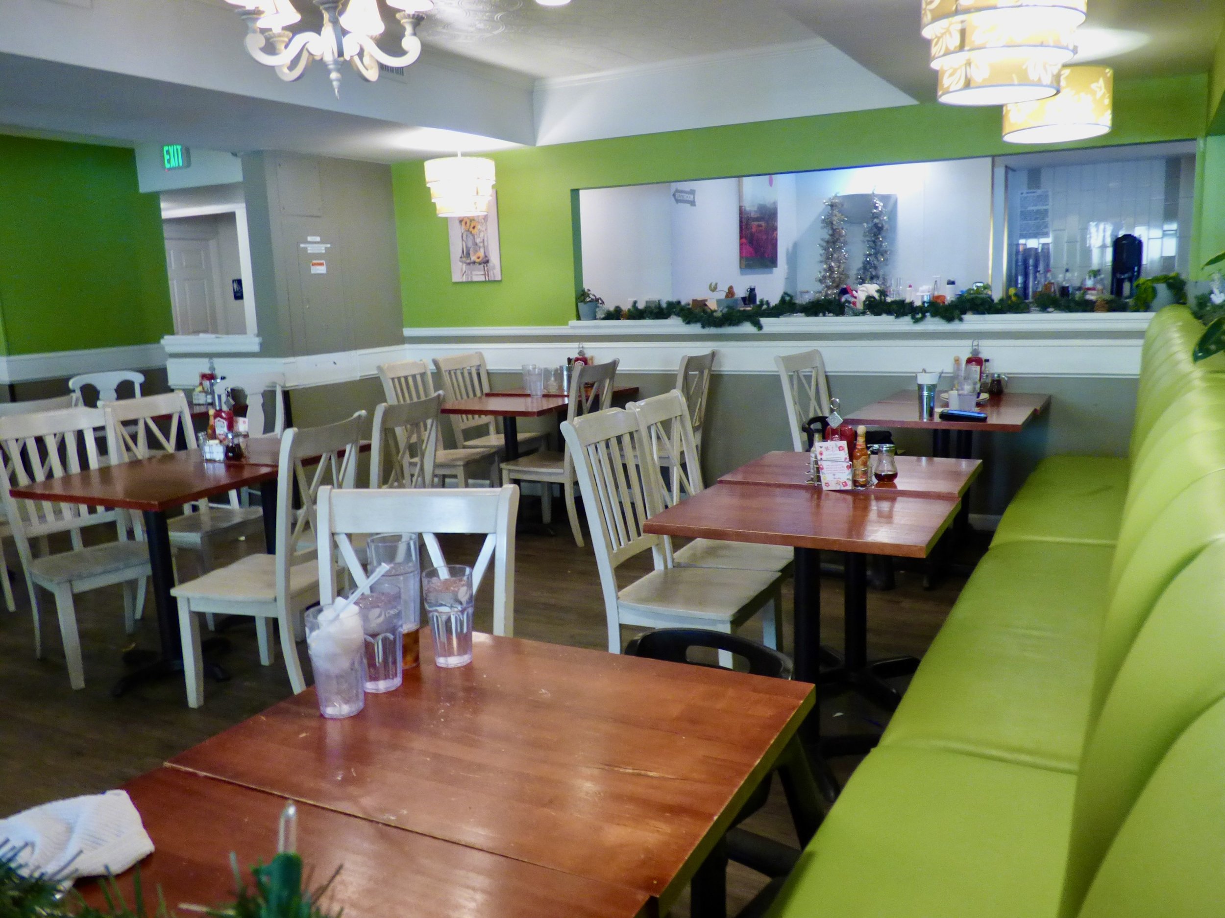 Empty restaurant with wooden tables and white chairs, lime green wall, and a holiday garland. Glasses with ice and straws on the tables, Christmas decorations visible through a window.