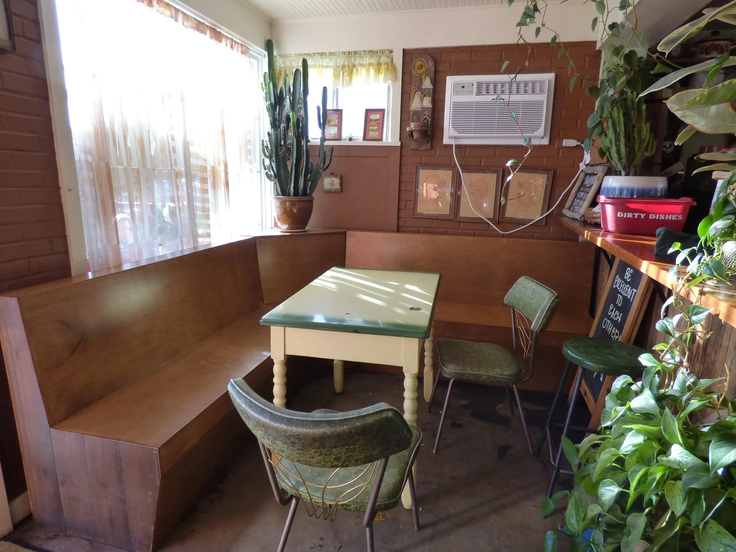 Cozy corner of a restaurant or cafe with a wooden booth seat, a small table, vintage green and brown chairs, potted plants, and a window with sheer curtains at Weathervane Cafe, Denver, CO.