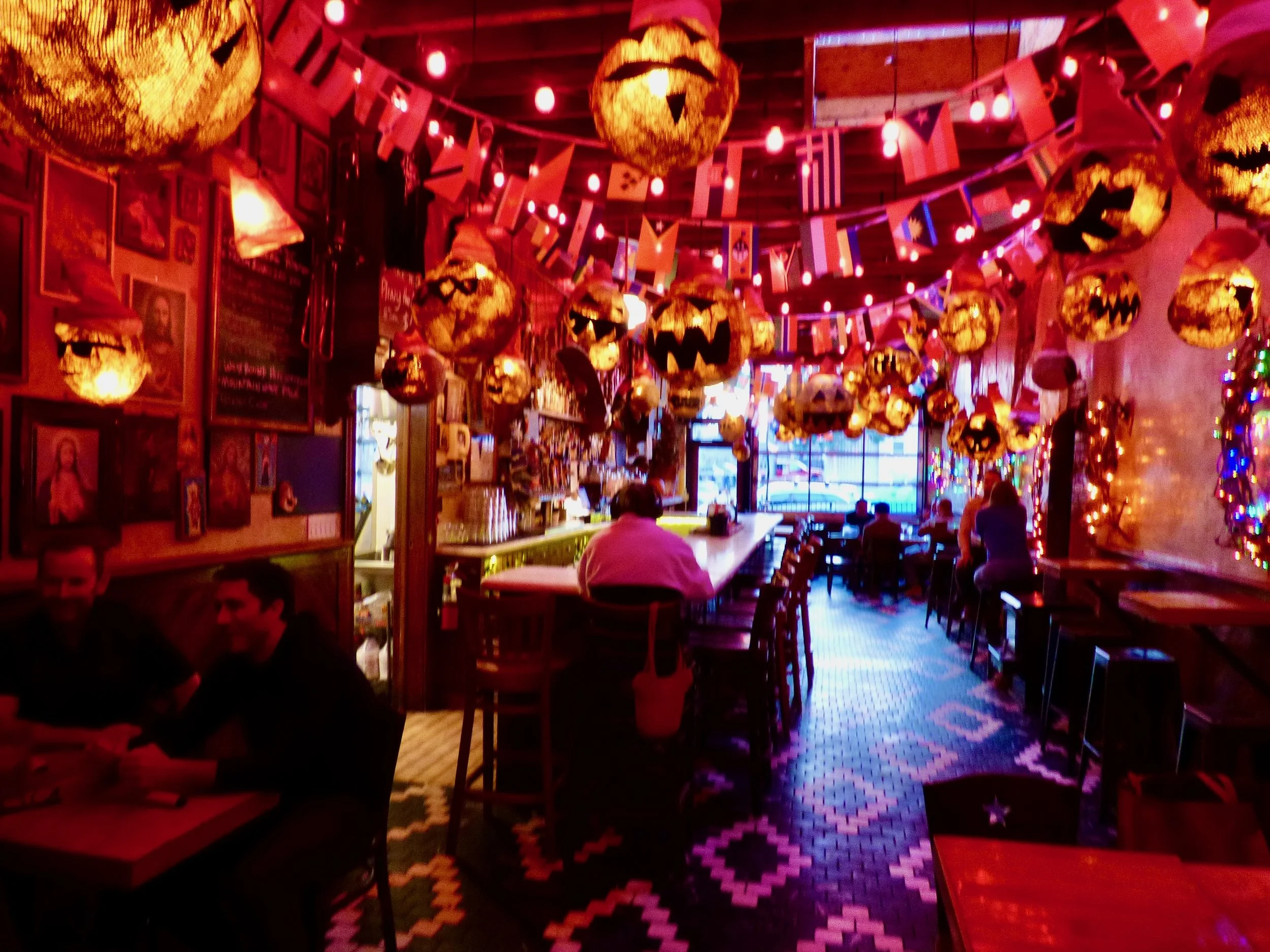 Interior of a restaurant decorated with Halloween themed paper lanterns and string lights, featuring flags hanging from the ceiling, with a few patrons sitting at the bar and tables at The Thin Man, Denver, CO.