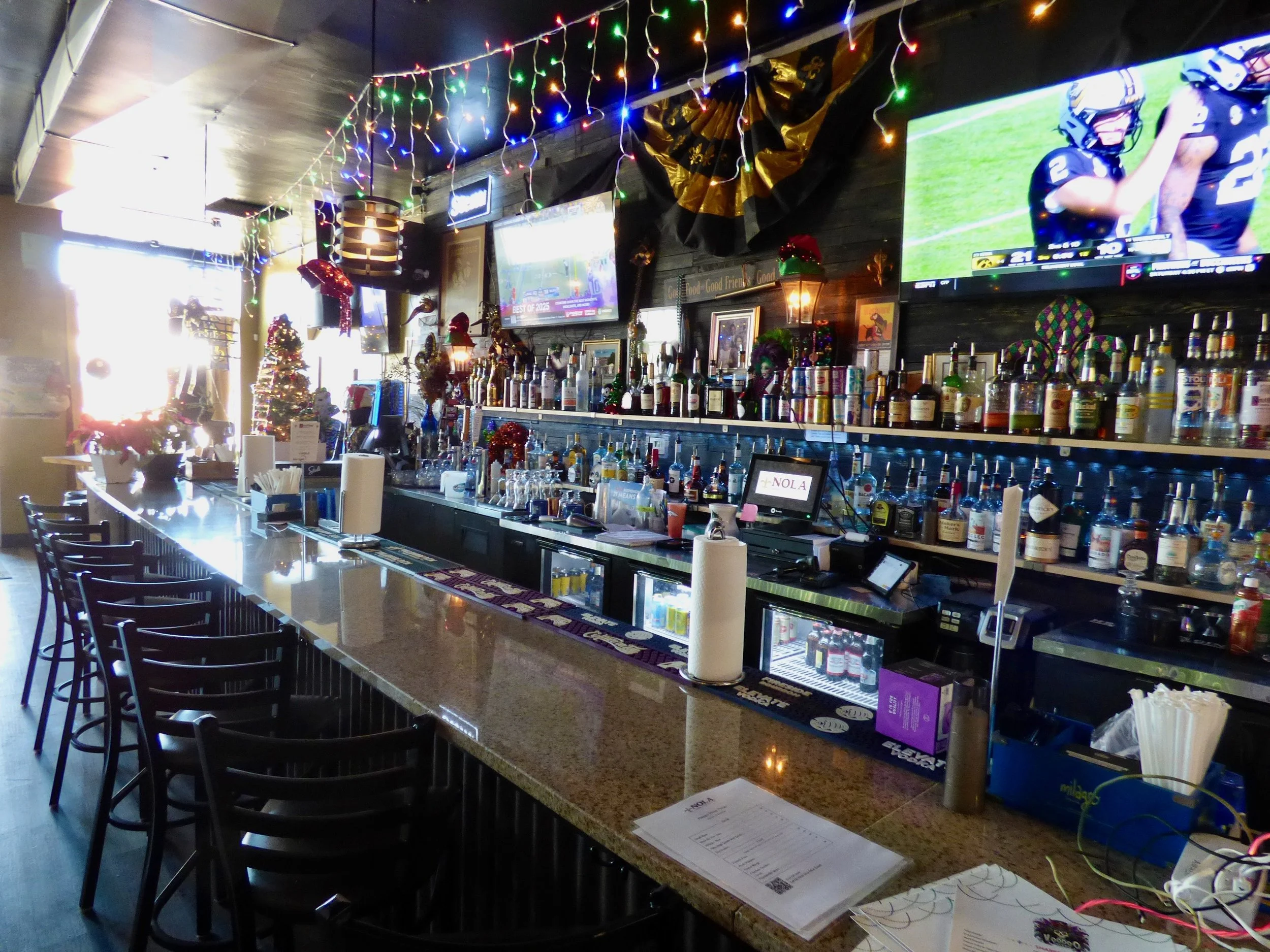 Interior of a bar decorated with Christmas and holiday decorations, including string lights, a small Christmas tree, and festive ornaments, with multiple TVs showing sports, a long granite bar counter at NOLA Voodoo Tavern, Denver, CO 80205.