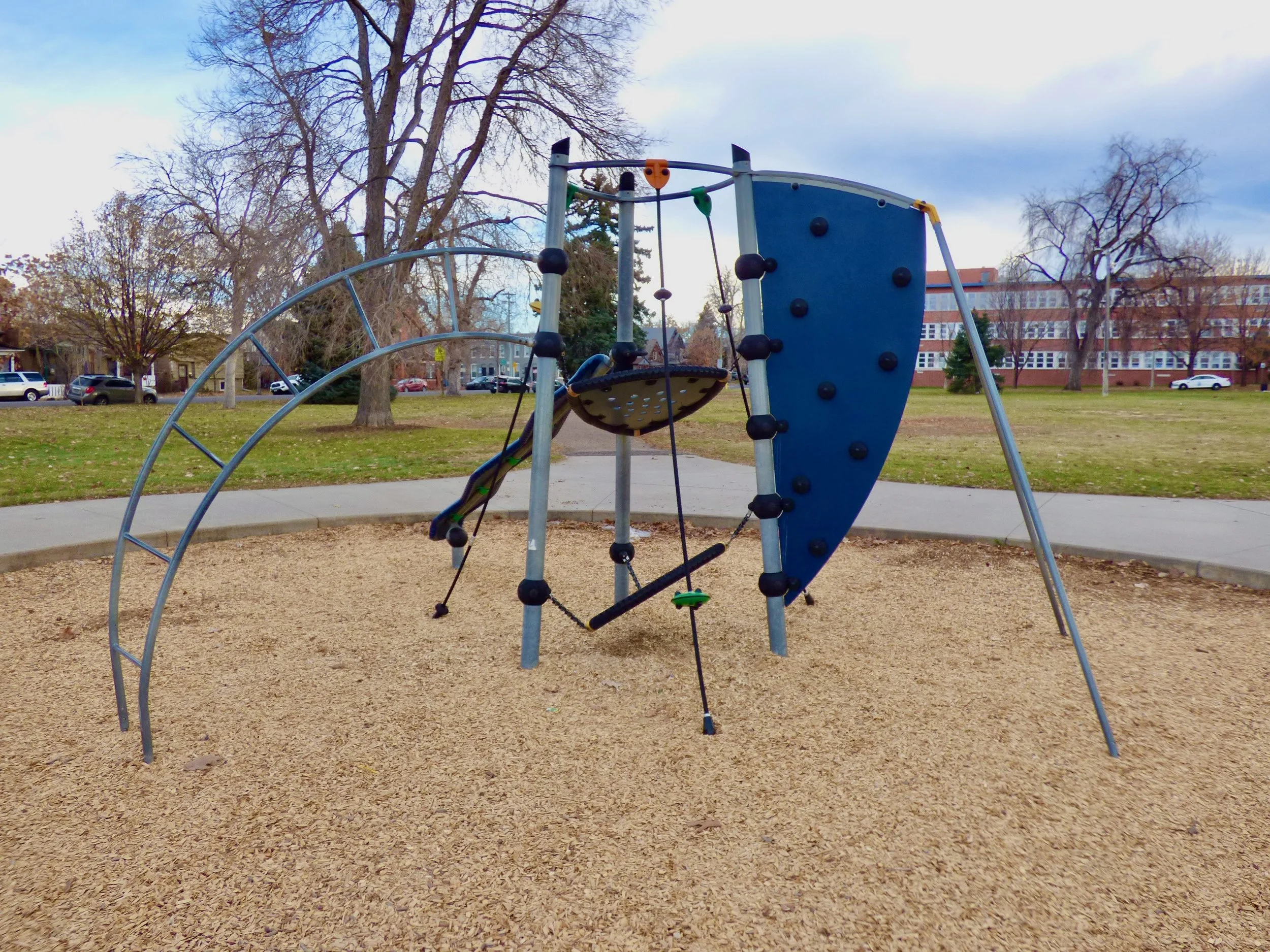 A children's climbing structure in a park on a gravel surface, with a rock climbing wall and a net climb, surrounded by trees and a sidewalk, with a school building in the background at Fuller Park, Denver, CO 80205.